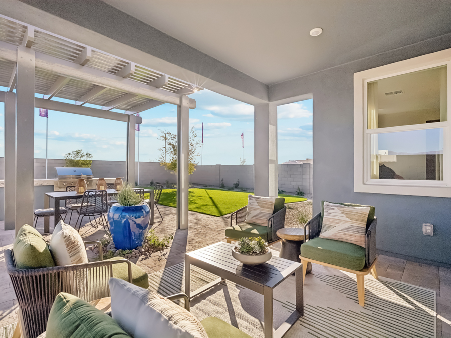 Modern covered patio with outdoor seating, dining table, potted plants, and a view of a green lawn and fenced backyard under a partly cloudy sky.