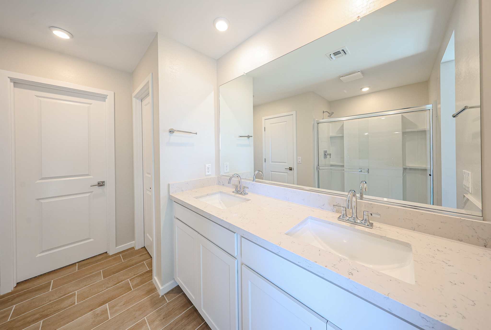 Modern bathroom with double sinks, large mirror, white cabinets, beige tile floor, and a glass-enclosed shower. Two doors are visible, one leading to another room and one to a closet or toilet.