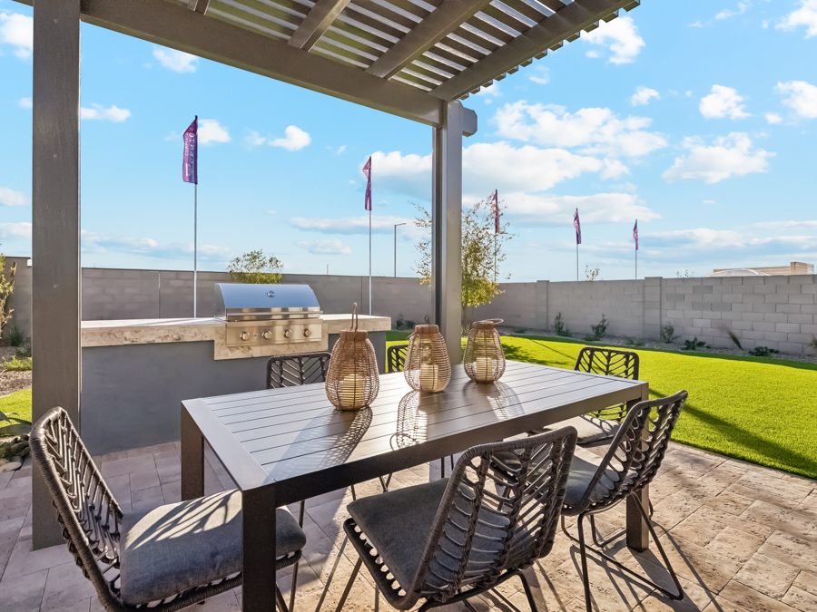 Outdoor patio with a pergola, dining table and chairs, decorative lanterns, built-in grill, and a grassy yard, enclosed by a block wall under a blue sky with scattered clouds.
