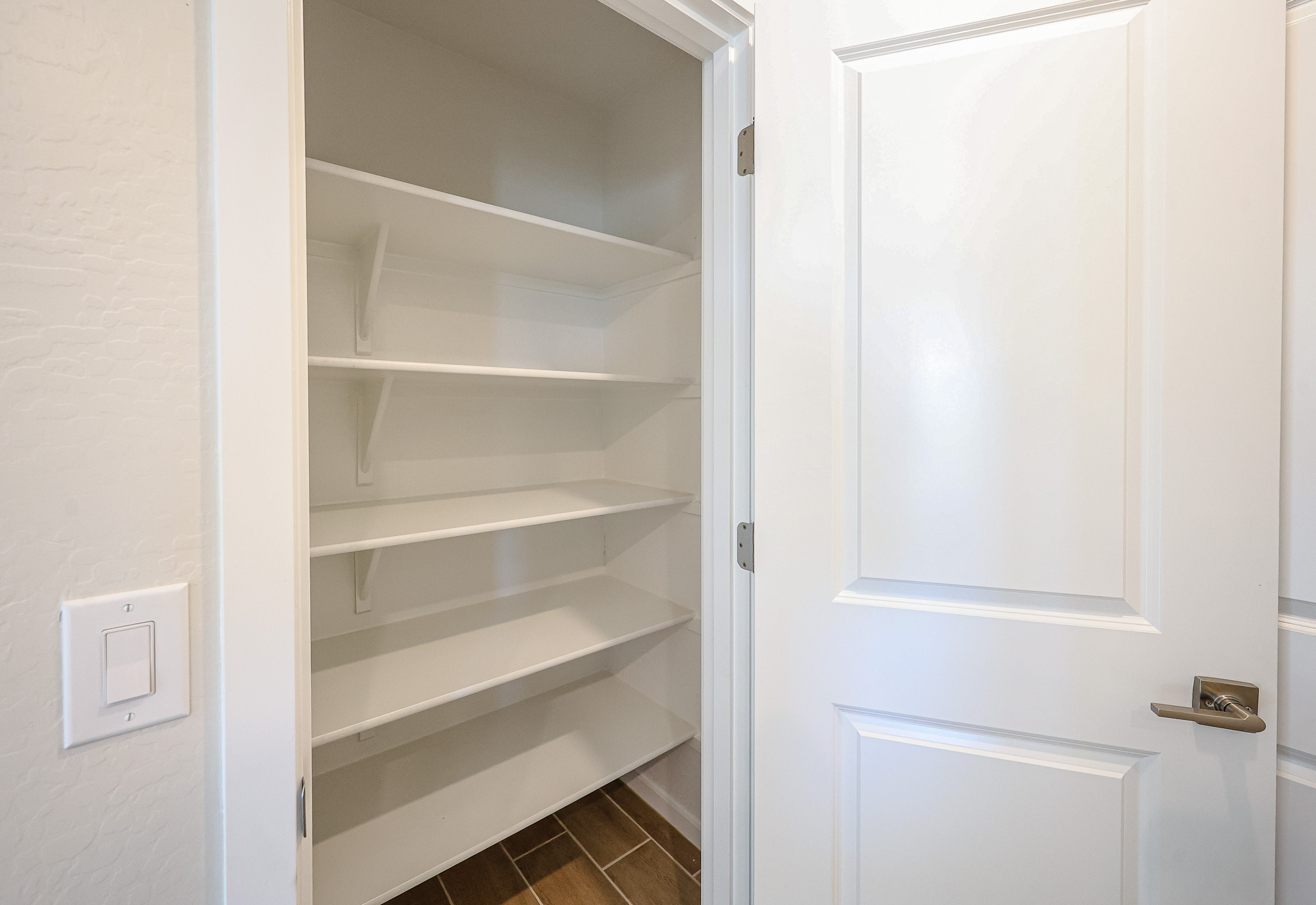 Open white door revealing an empty pantry with four white shelves, white walls, brown tile floor, and a light switch on the wall outside.