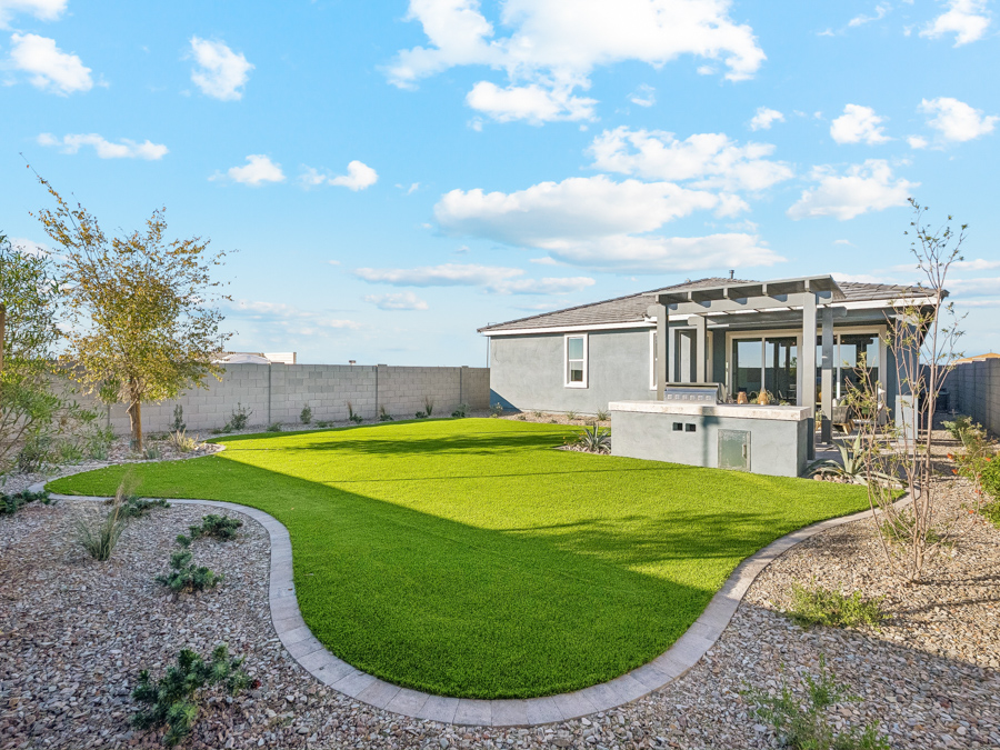 Backyard with green grass lawn, rock landscaping, and a modern gray house with a covered patio under a partly cloudy sky.