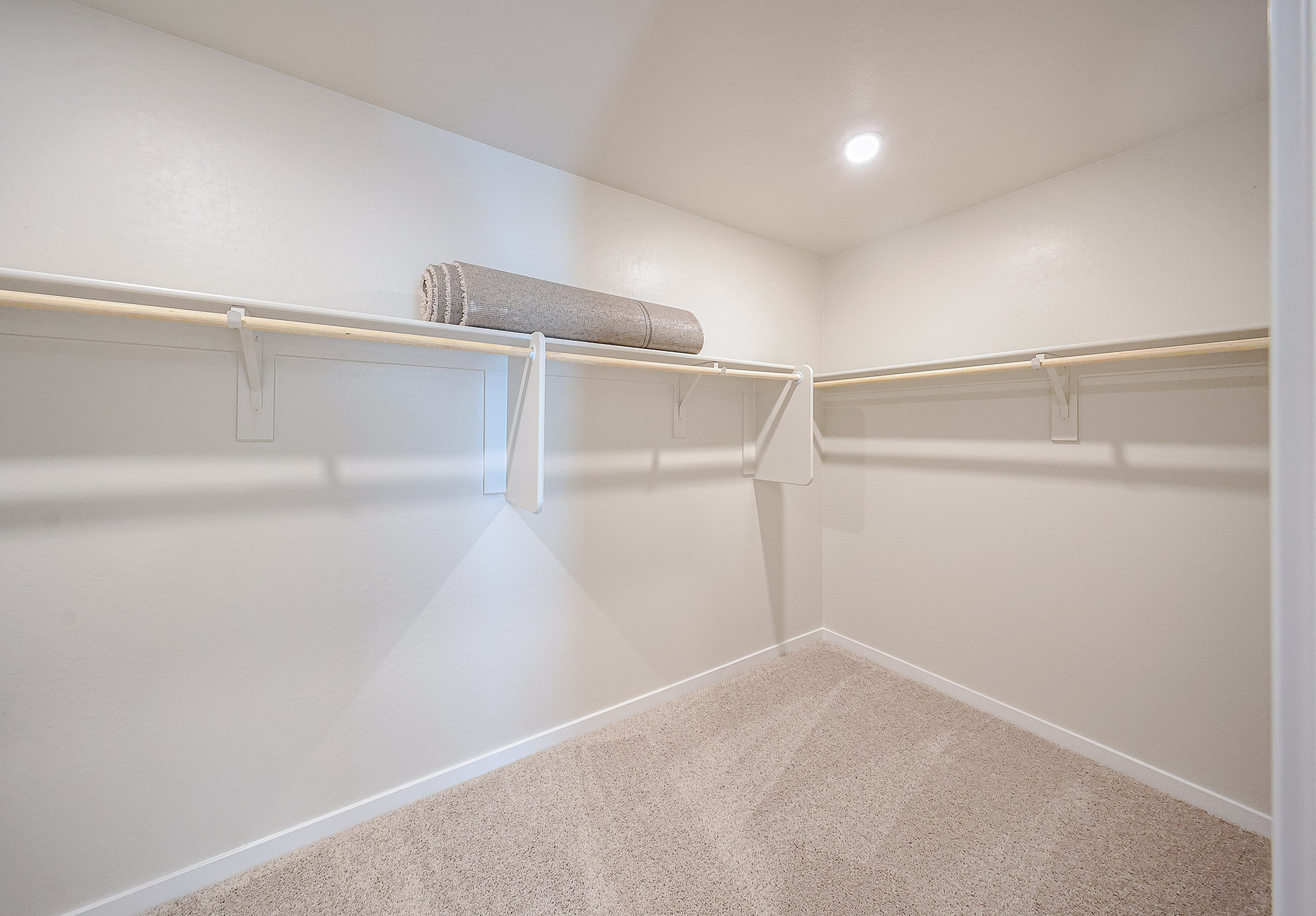 Empty walk-in closet with beige carpet, white walls, two white shelves with hanging rods, and a rolled-up gray rug on the top shelf, lit by a ceiling light.