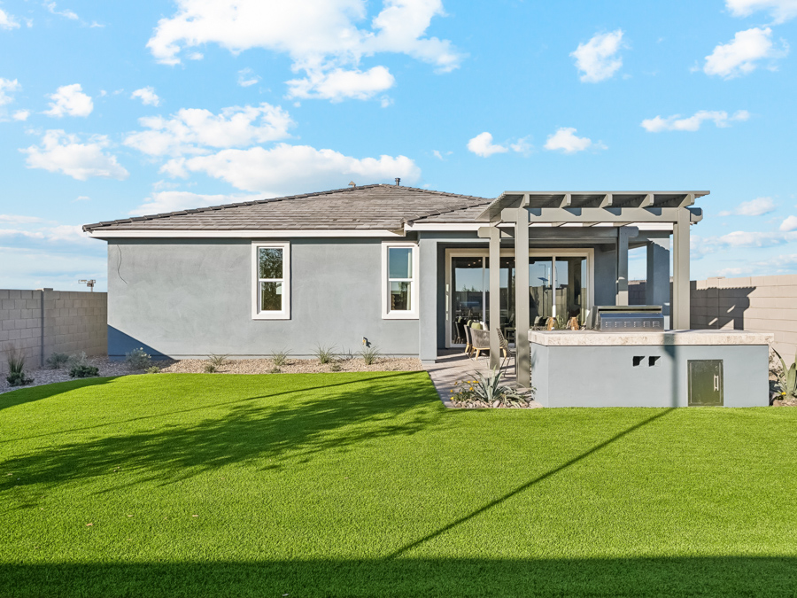 Single-story gray house with a patio, pergola, and outdoor kitchen, surrounded by a green lawn and enclosed by a beige block wall under a blue sky with clouds.