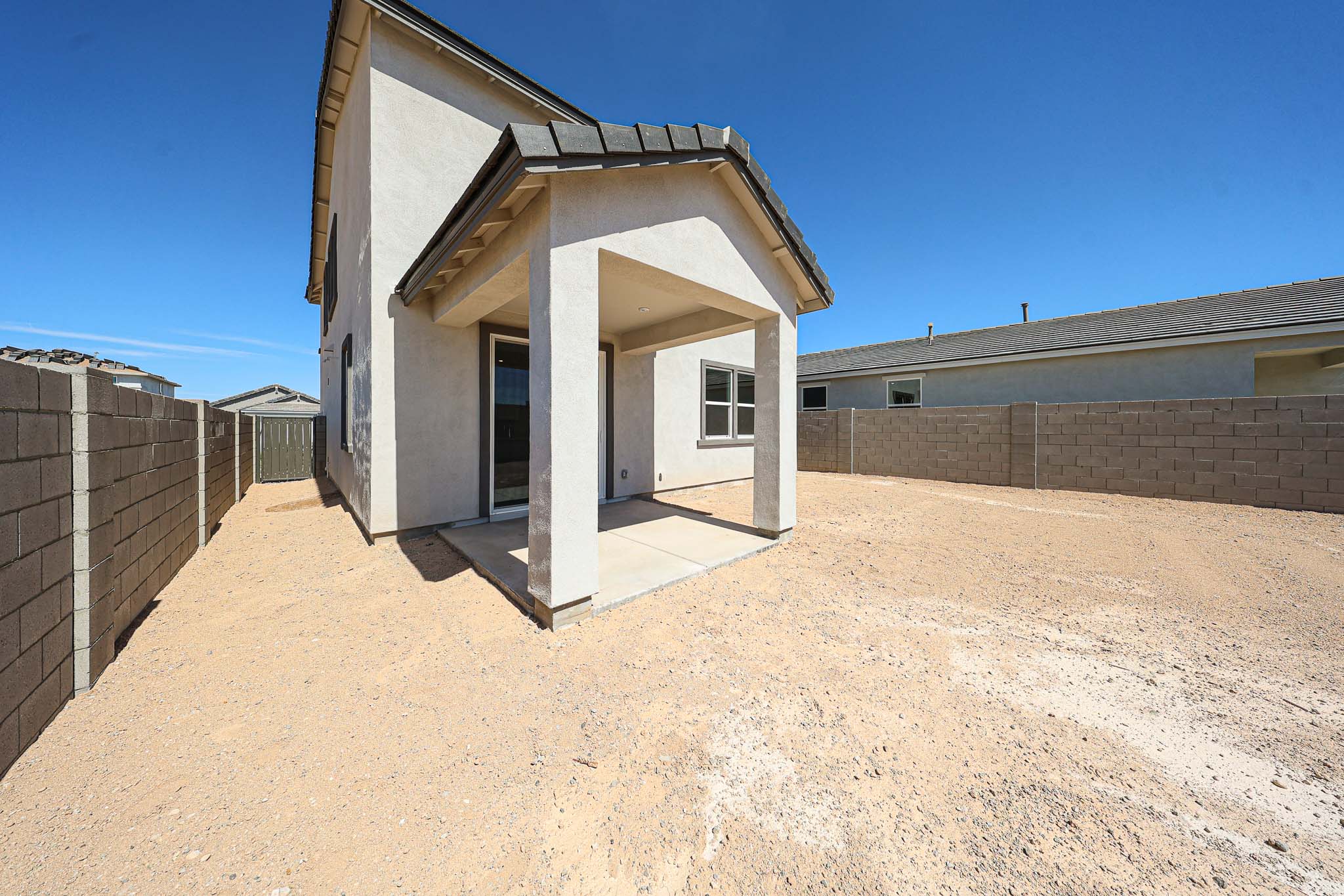 Two-story house with a covered patio and fenced backyard, surrounded by gravel and concrete block walls under a clear blue sky.