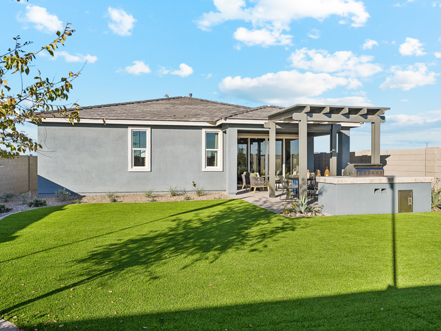 A modern single-story house with gray exterior, covered patio, outdoor seating, grill area, and a large green lawn under a blue sky with clouds.