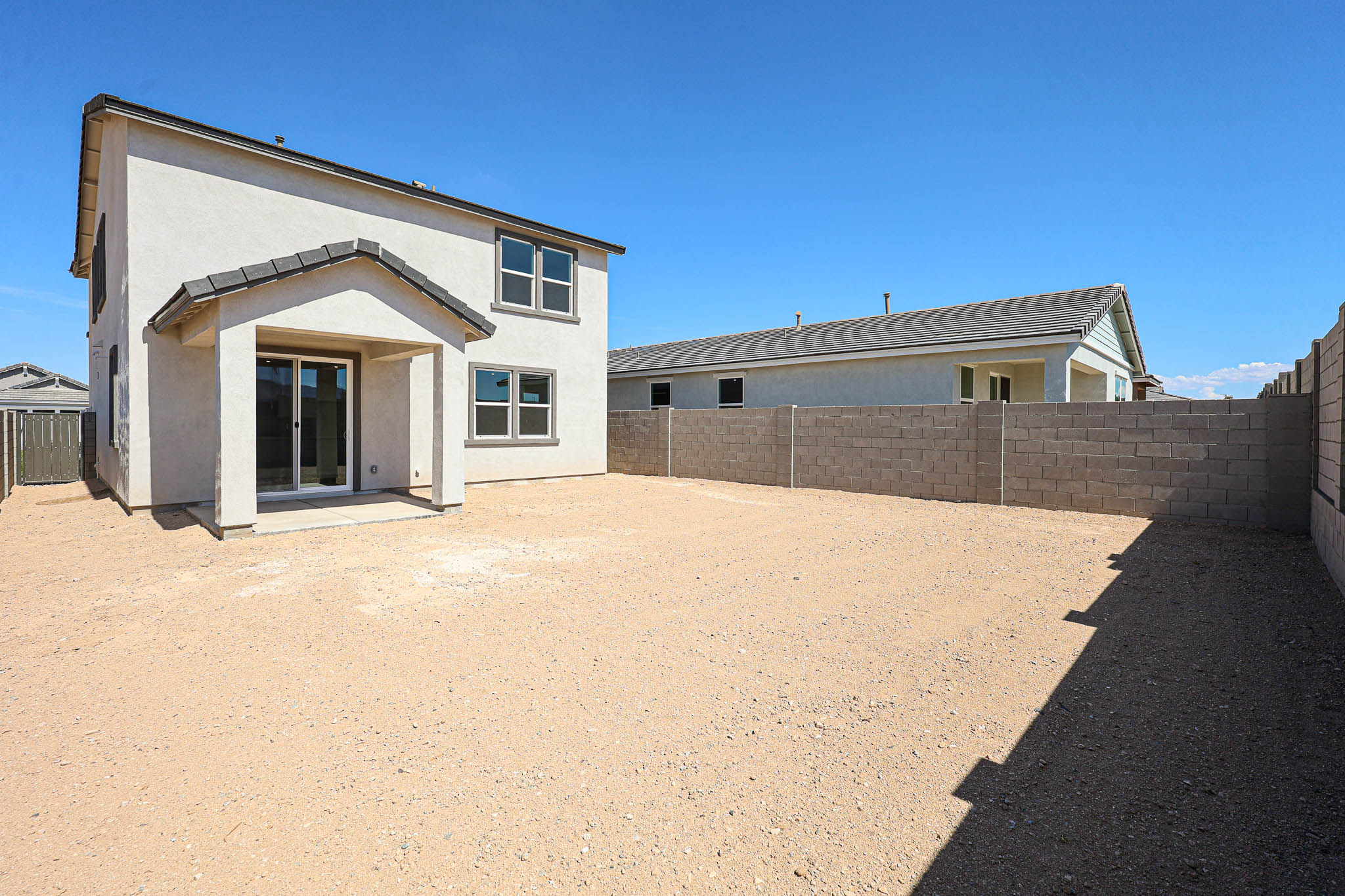 A two-story house with a covered back patio and large, unfurnished dirt backyard enclosed by a block wall under a clear blue sky.