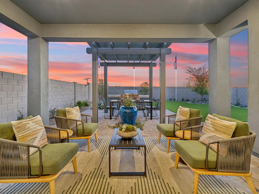 Covered patio with four green cushioned chairs, a coffee table, and a pergola-covered dining area in a fenced backyard at sunset.