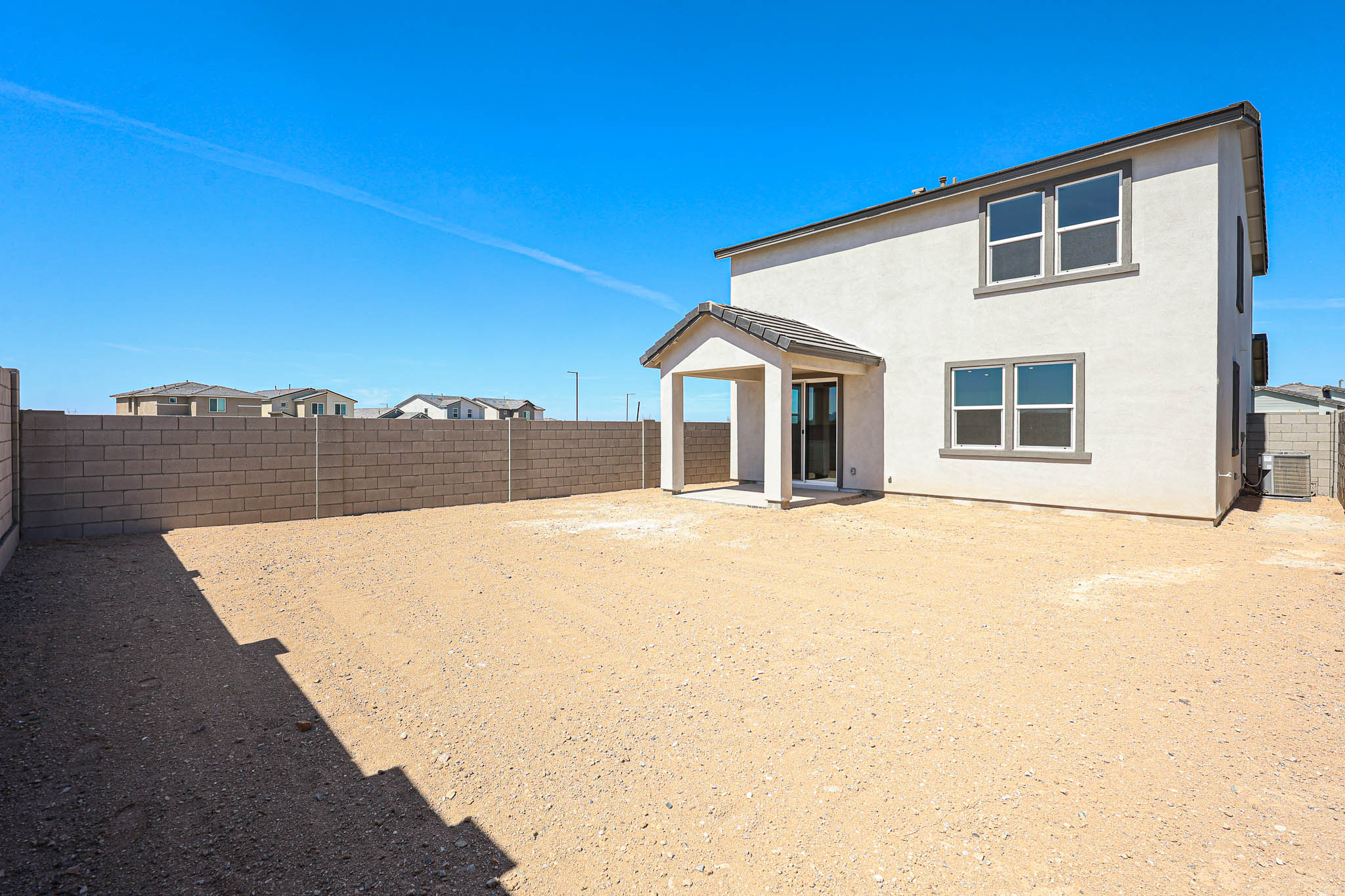 Two-story house with light-colored exterior, covered patio, and large unfurnished backyard enclosed by a block wall, under a clear blue sky.