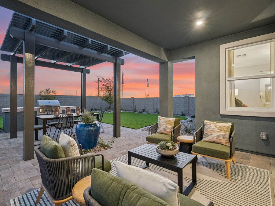 Covered patio with modern outdoor seating, a dining table under a pergola, potted plants, and a grassy yard at sunset.