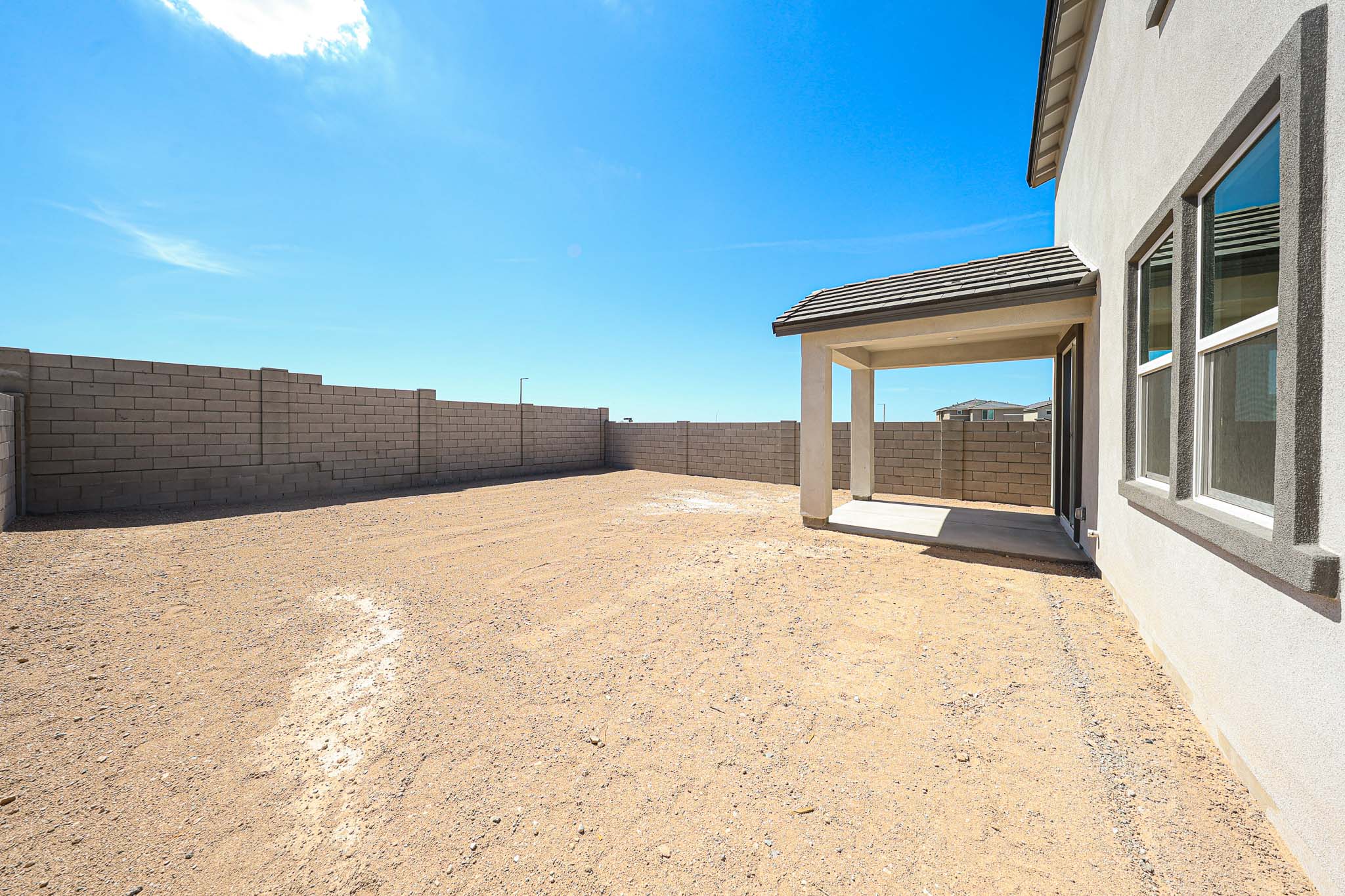 A large backyard with bare dirt, surrounded by a tall concrete block wall, and a covered patio attached to a modern house under a clear blue sky.