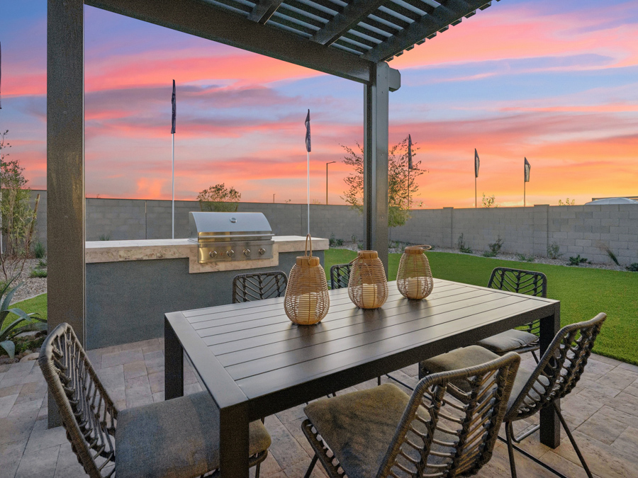 Outdoor patio with a black dining table, six chairs, wicker lanterns, a built-in grill, and a pergola, set against a sunset sky and fenced backyard with grass.