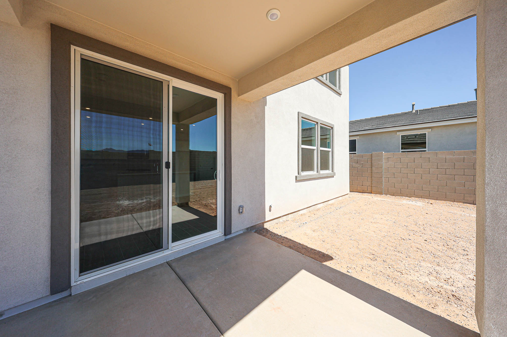 Covered patio with sliding glass door opens to a bare dirt backyard enclosed by a concrete block wall; neighboring house is visible under clear blue sky.