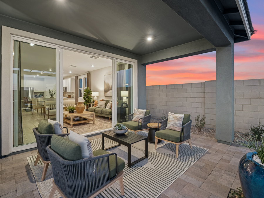 Modern covered patio with four chairs and a table on a rug, overlooking a living room through large sliding glass doors at sunset.