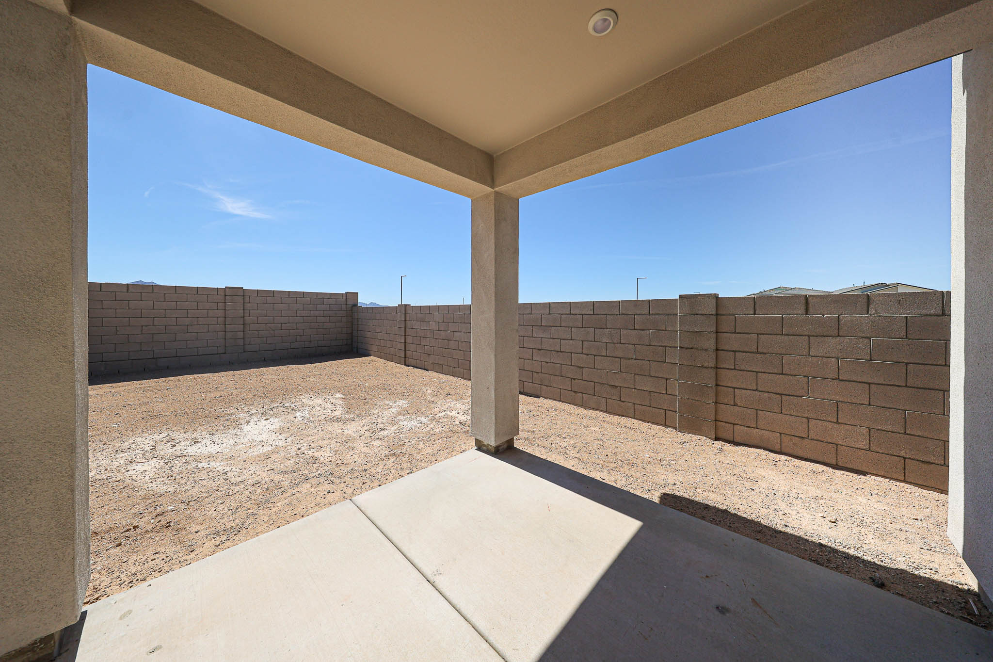View from a covered patio looking out onto a bare backyard enclosed by a tall cinder block wall under a clear blue sky.