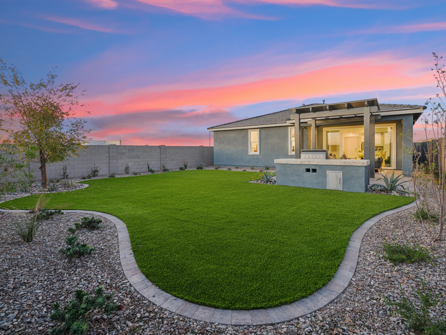 A modern backyard with artificial grass, a gravel border, small plants, and a covered patio at sunset.