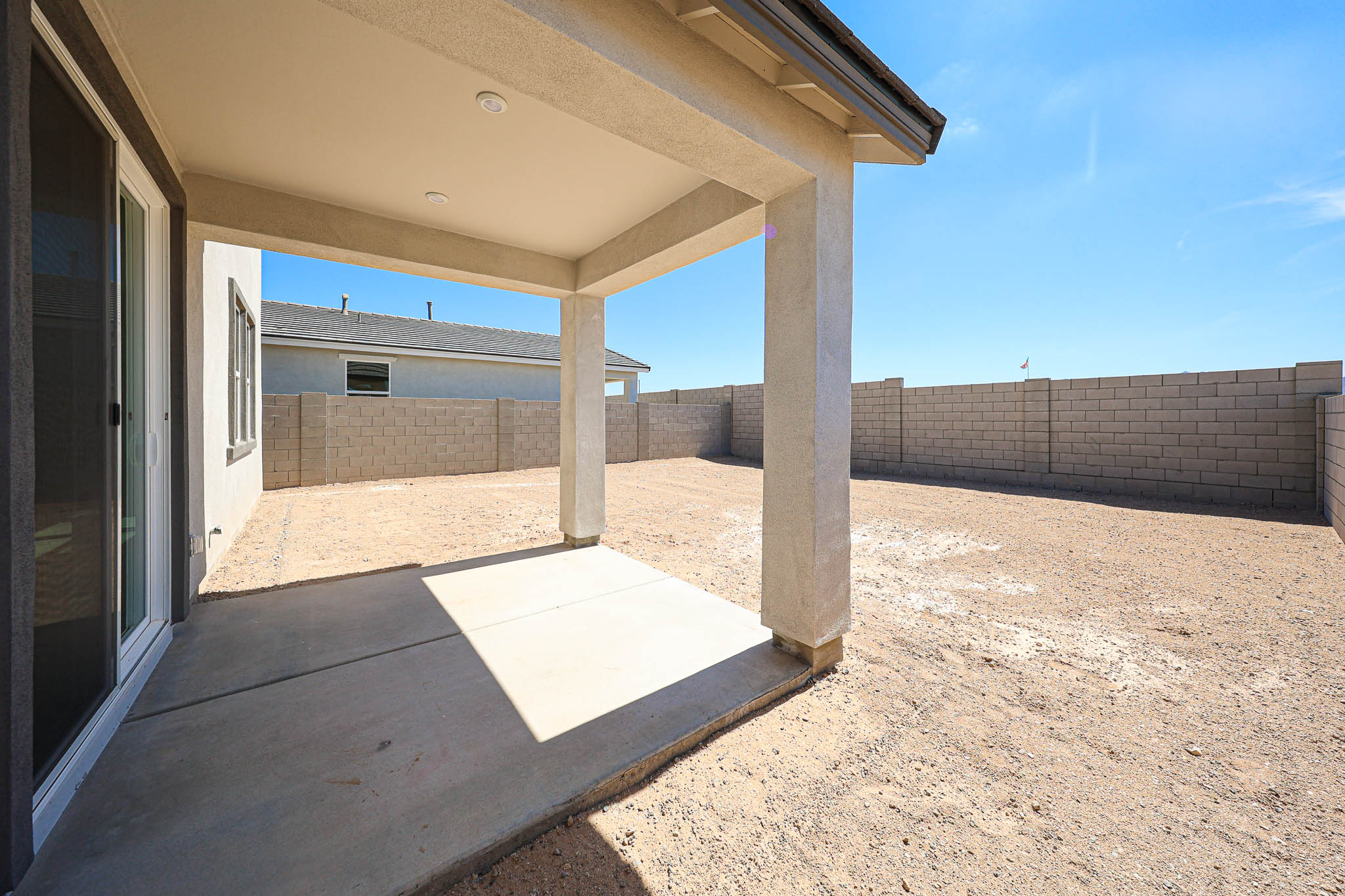 Covered concrete patio attached to a house, overlooking a large fenced backyard with bare dirt and a block wall under a clear blue sky.