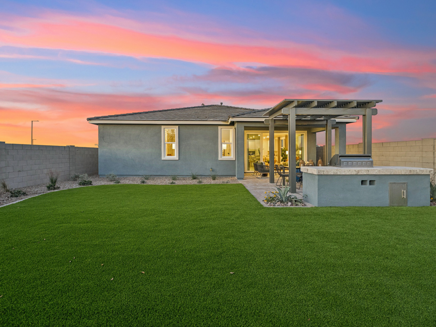 Single-story house with gray exterior, covered patio, built-in grill, and a large artificial turf backyard, under a colorful sunset sky.