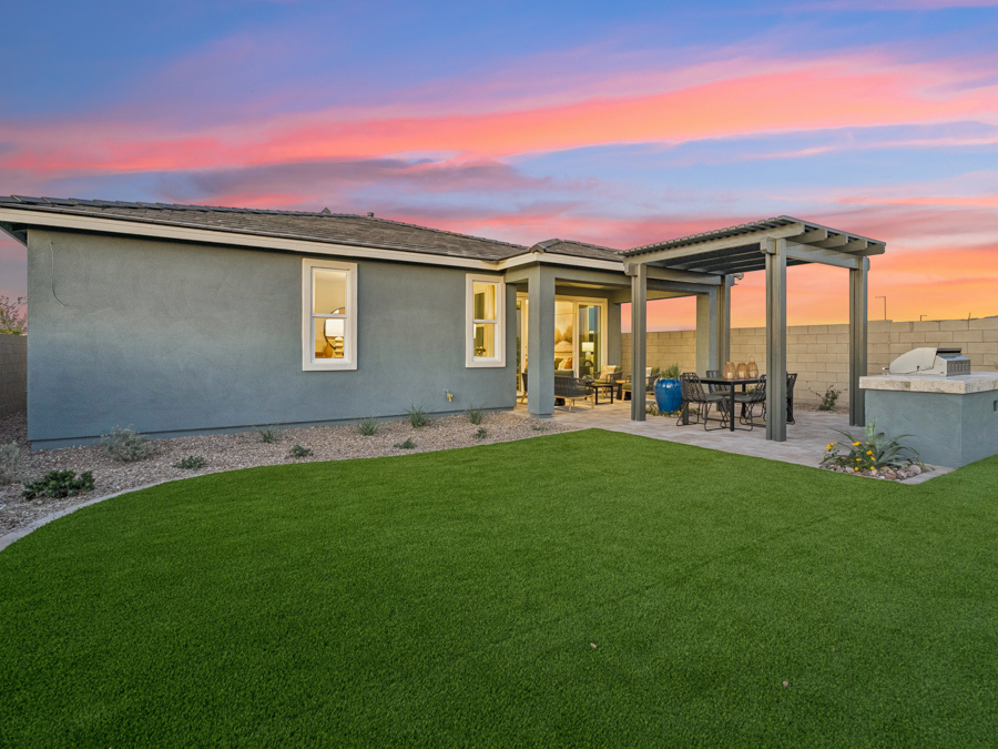 A modern single-story house with a covered patio, outdoor dining area, built-in grill, and artificial turf yard at sunset.