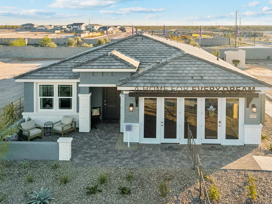 Single-story modern house with a tiled roof, large front windows, patio furniture, and a sign reading “A HOME FOR EVERY DREAM” above the entrance. Desert landscaping surrounds the property.