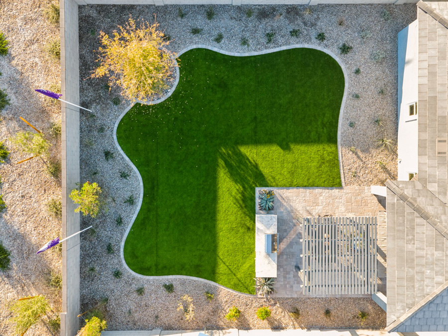 Aerial view of a backyard with a curved patch of green grass, stone patio, pergola, and surrounding gravel landscaping.