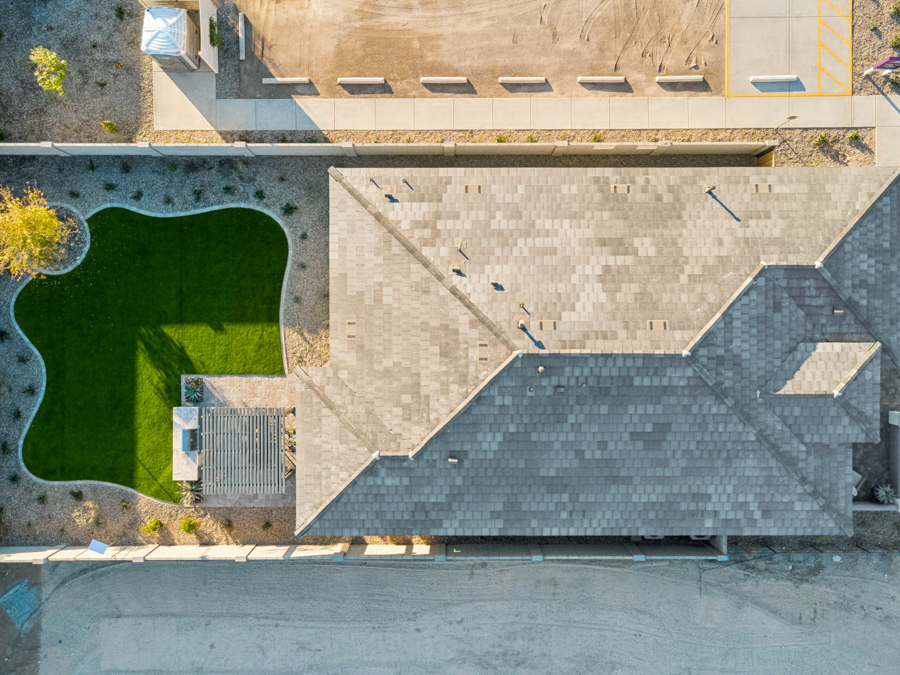 Aerial view of a house with a gray shingle roof, adjacent green lawn with landscaping, and surrounding gravel areas.