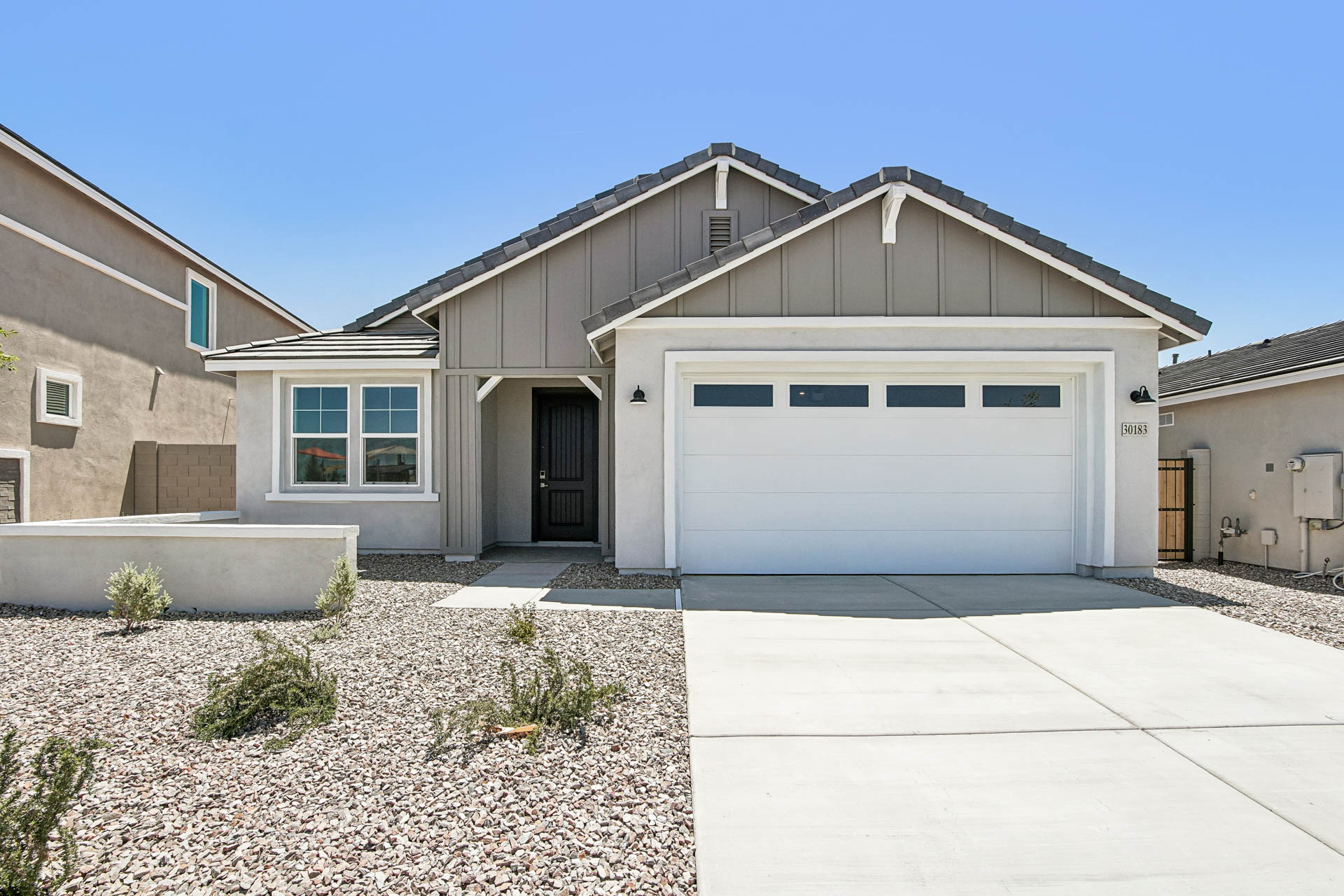 Single-story modern house with gray siding, white trim, a two-car garage, and a gravel yard under a clear blue sky.