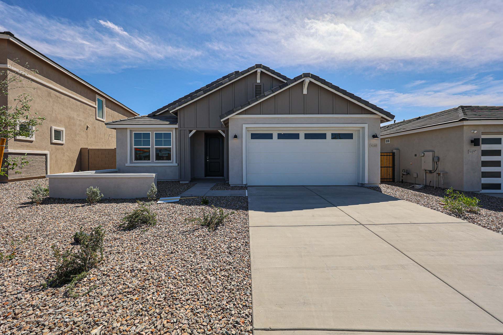 Single-story house with a two-car garage, beige exterior, and gravel landscaping in front, under a partly cloudy sky.
