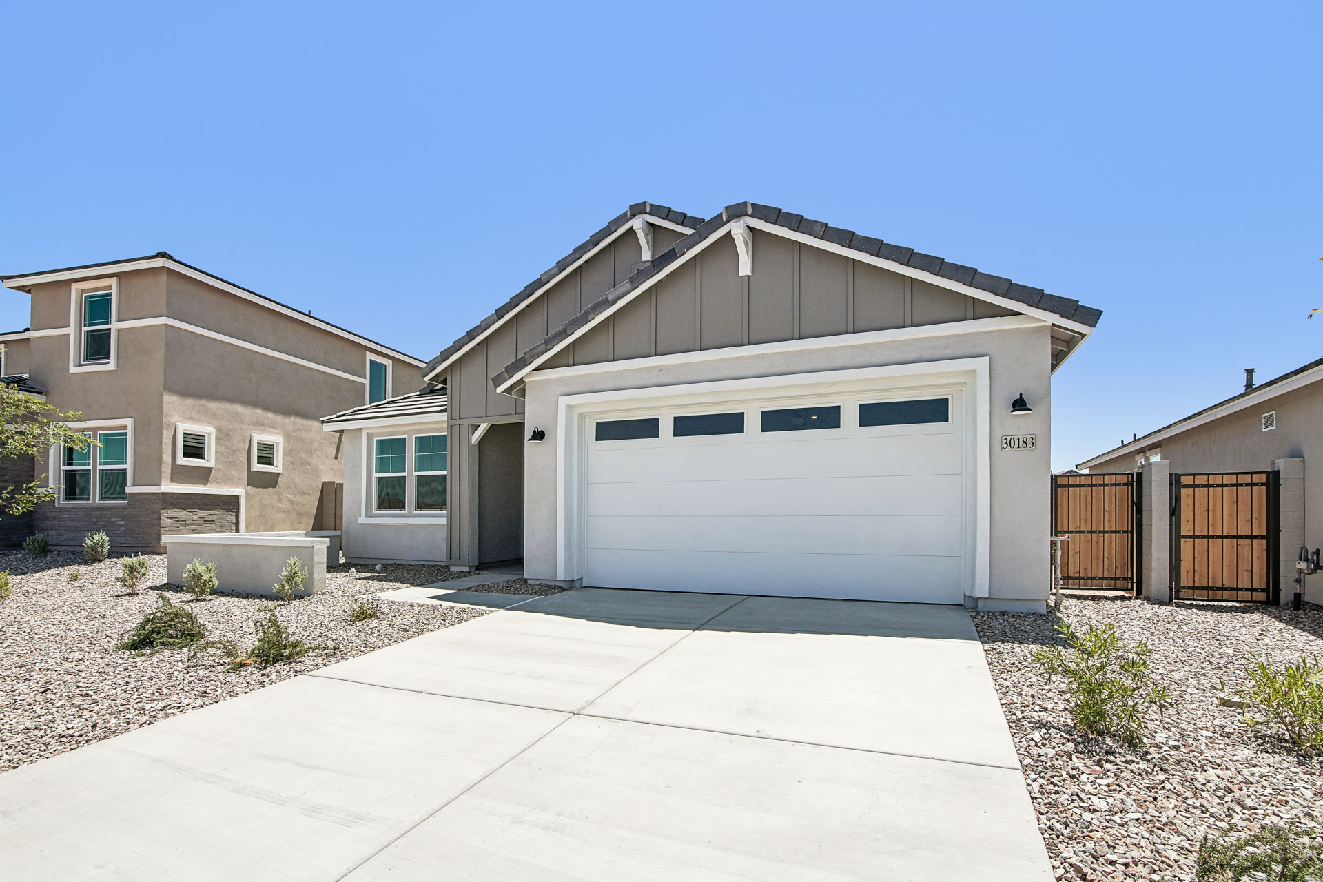 Single-story modern house with gray and white exterior, attached two-car garage, and low-maintenance desert landscaping under a clear blue sky.