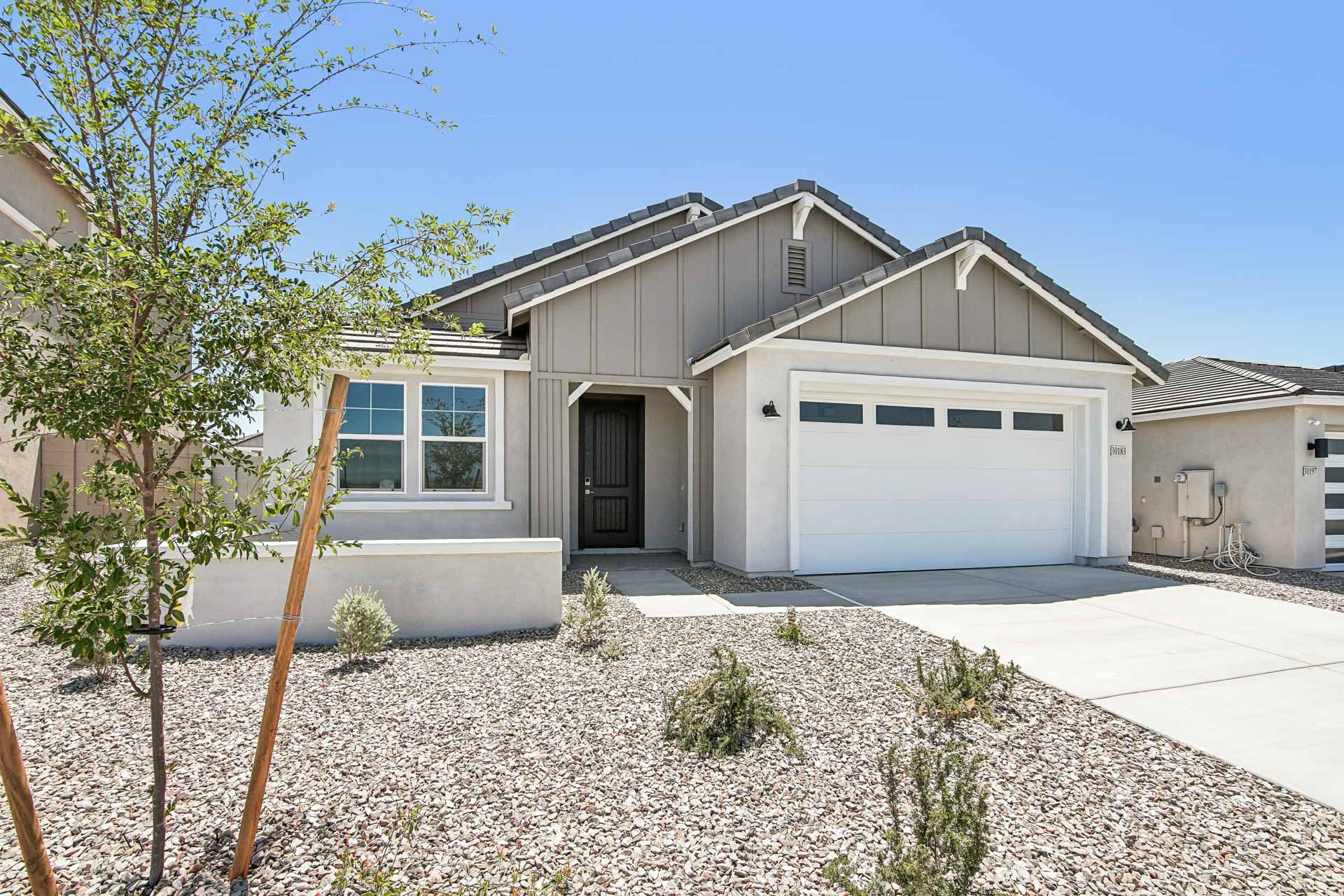 Single-story modern house with gray siding, white trim, and a two-car garage. Xeriscaped front yard with rocks, small shrubs, and a tree. Clear sky and neighboring houses visible.