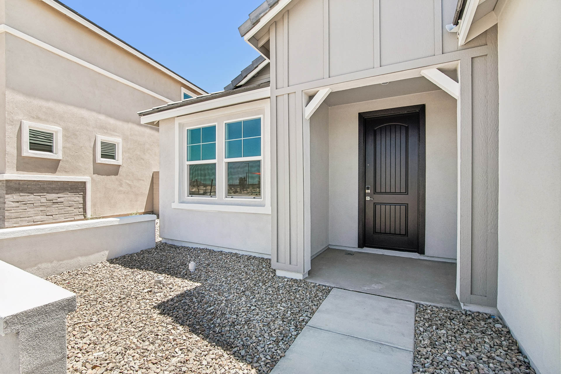 Modern house entrance with white walls, a covered porch, dark front door, and a gravel yard under bright sunlight.