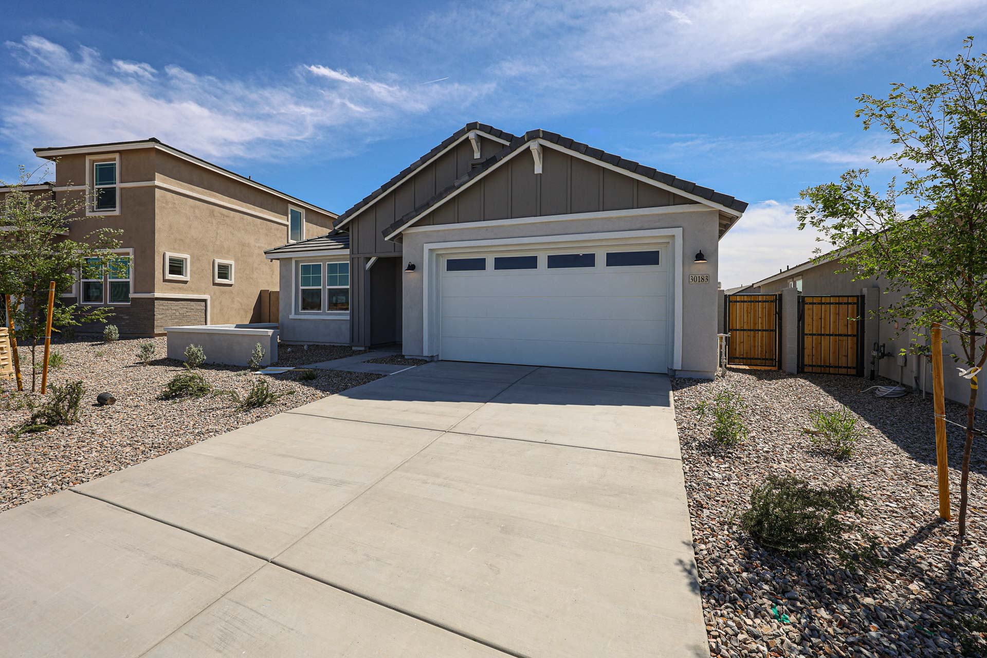 Single-story house with a gray facade, white garage door, and a concrete driveway, surrounded by low-maintenance desert landscaping under a blue sky.