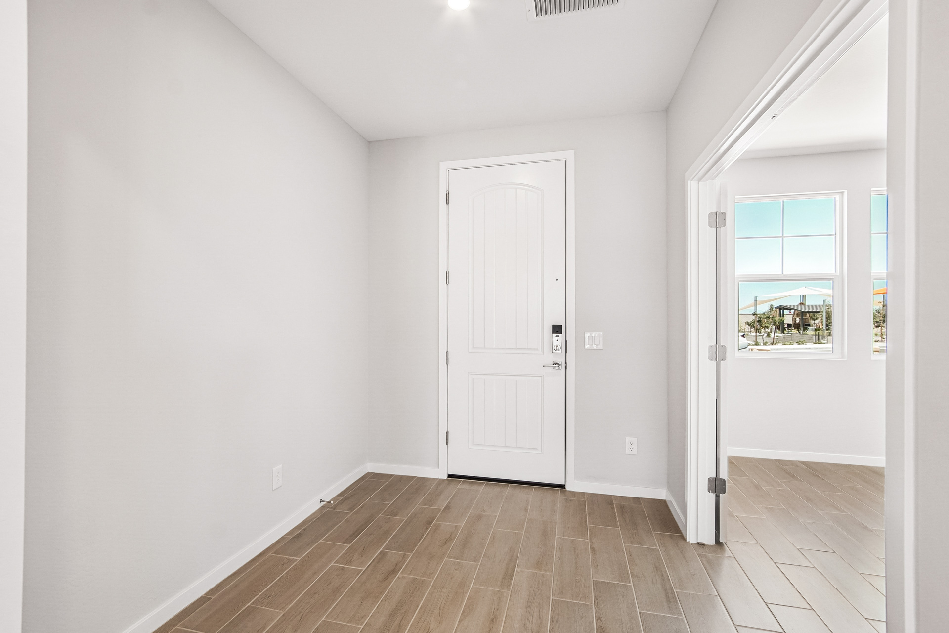 A clean, empty hallway with light-colored walls, a wooden floor, a closed front door, and an open doorway leading to a bright room with windows.
