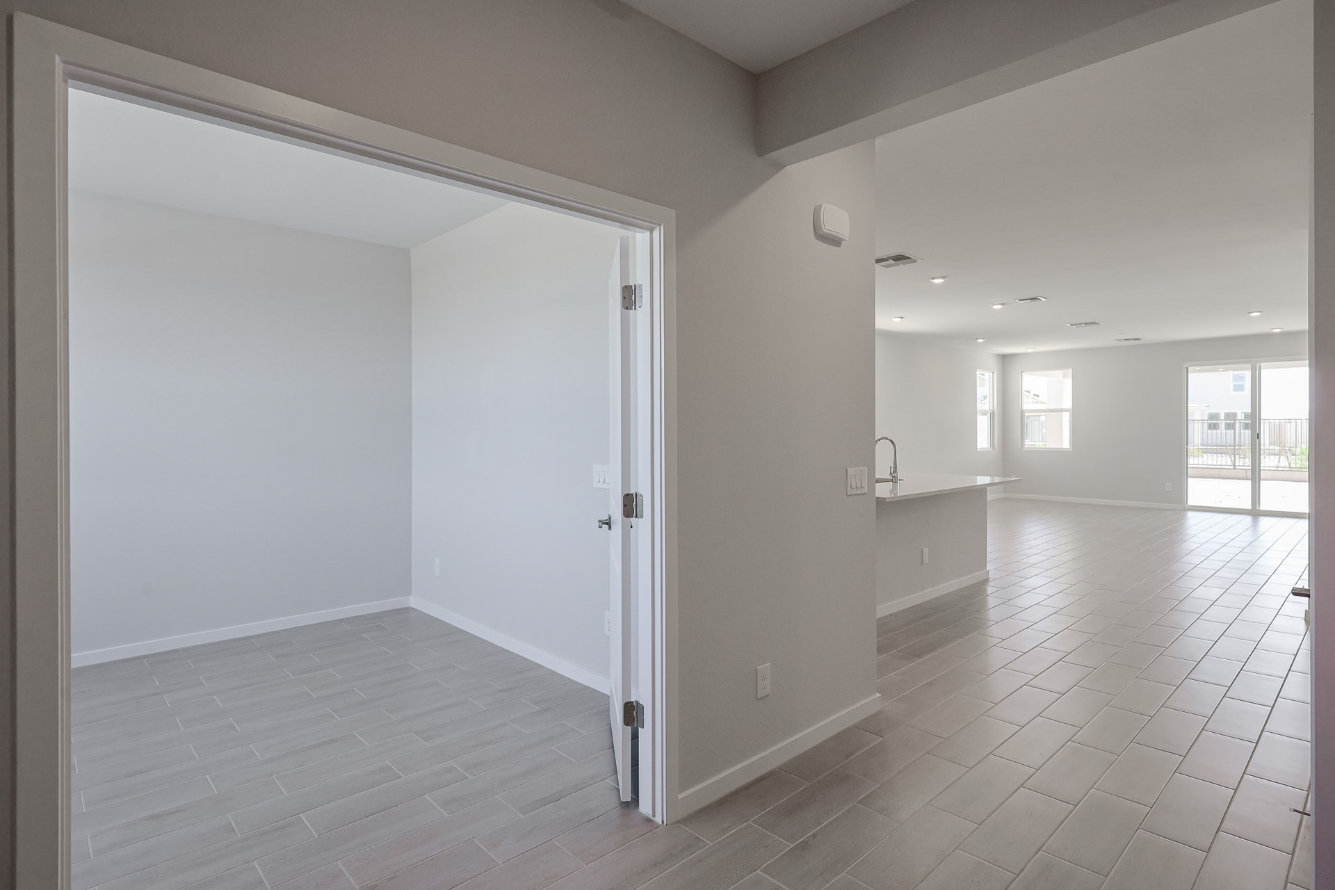 A modern, unfurnished home interior with tiled floors, an empty room on the left, and an open-plan kitchen and living area on the right.