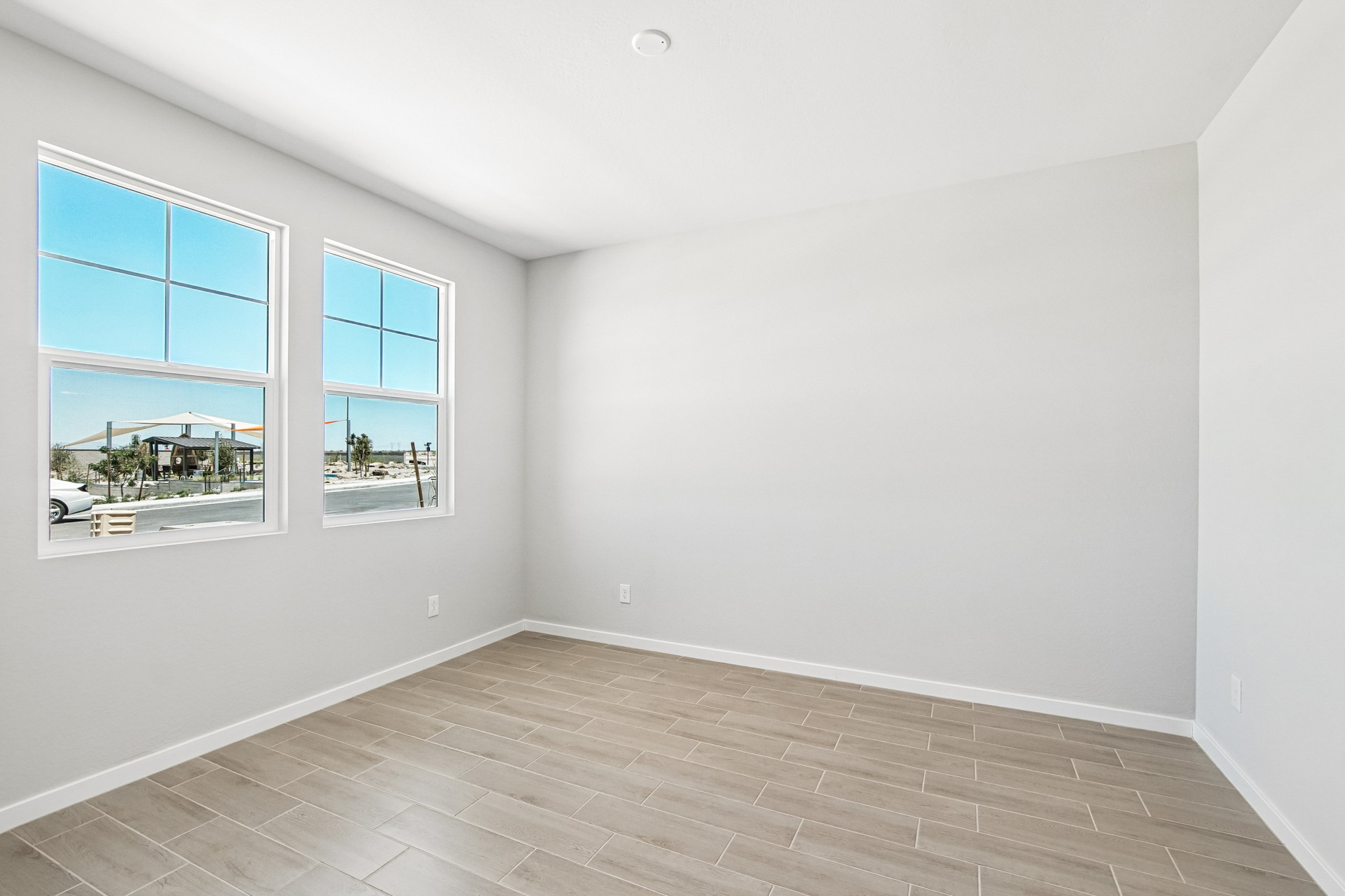 Empty room with light gray walls, two large windows, beige wood-look tile flooring, and white ceiling. Daylight streams in through the windows, showing a street view outside.