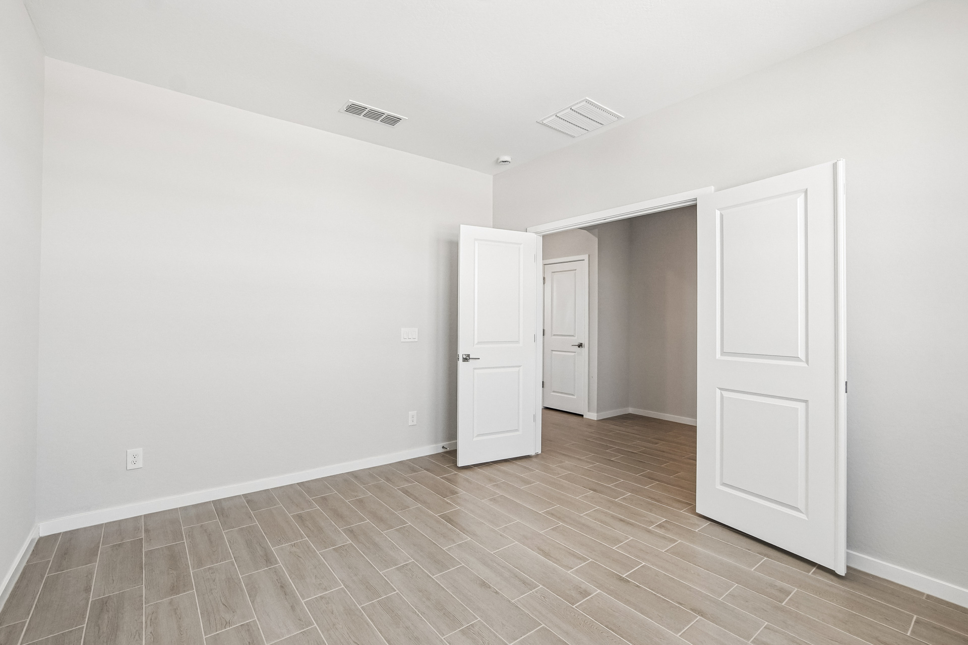 Empty room with light-colored tile flooring, white walls, and three open white doors leading to adjacent spaces. Ceiling vents and a light switch are visible.