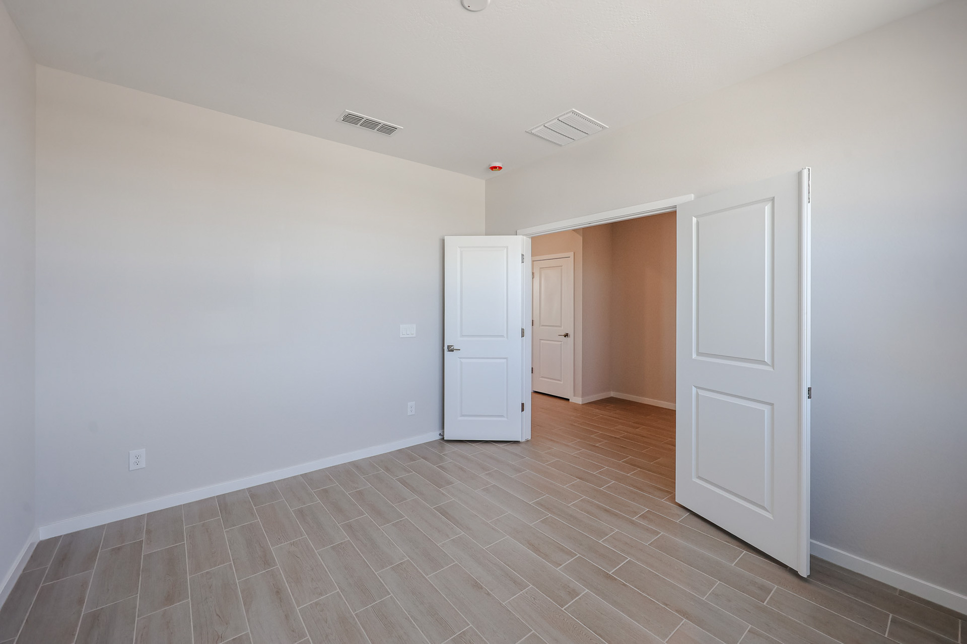 Empty room with beige tile flooring, white walls, and open white double doors leading to another hallway and door. Ceiling vents and a red smoke detector are visible.