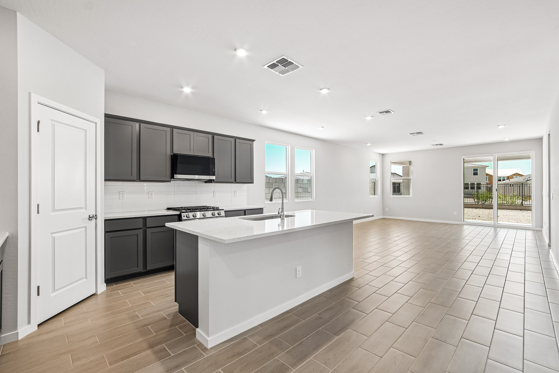 Modern open-concept kitchen and living area with gray cabinets, white countertops, tile flooring, and large windows offering natural light and a view of the backyard.