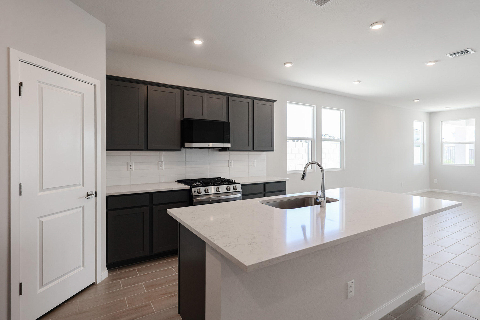 Modern kitchen with dark cabinets, stainless steel appliances, a center island with sink, light countertops, white backsplash, and large windows letting in natural light.