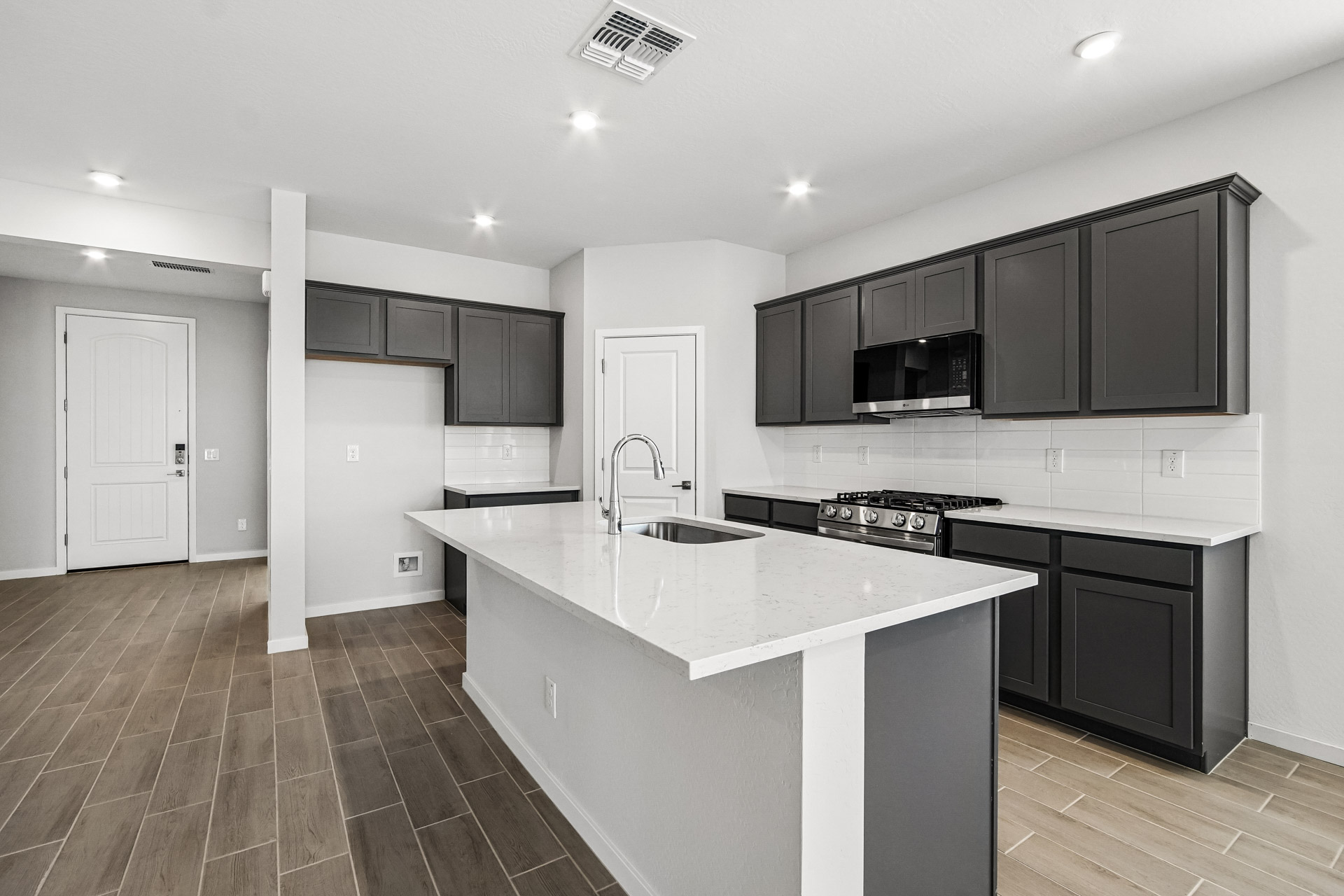 Modern kitchen with dark cabinets, white countertops, stainless steel appliances, and wood-look tile flooring. A central island features a sink and faucet. Entryway and empty space are visible.