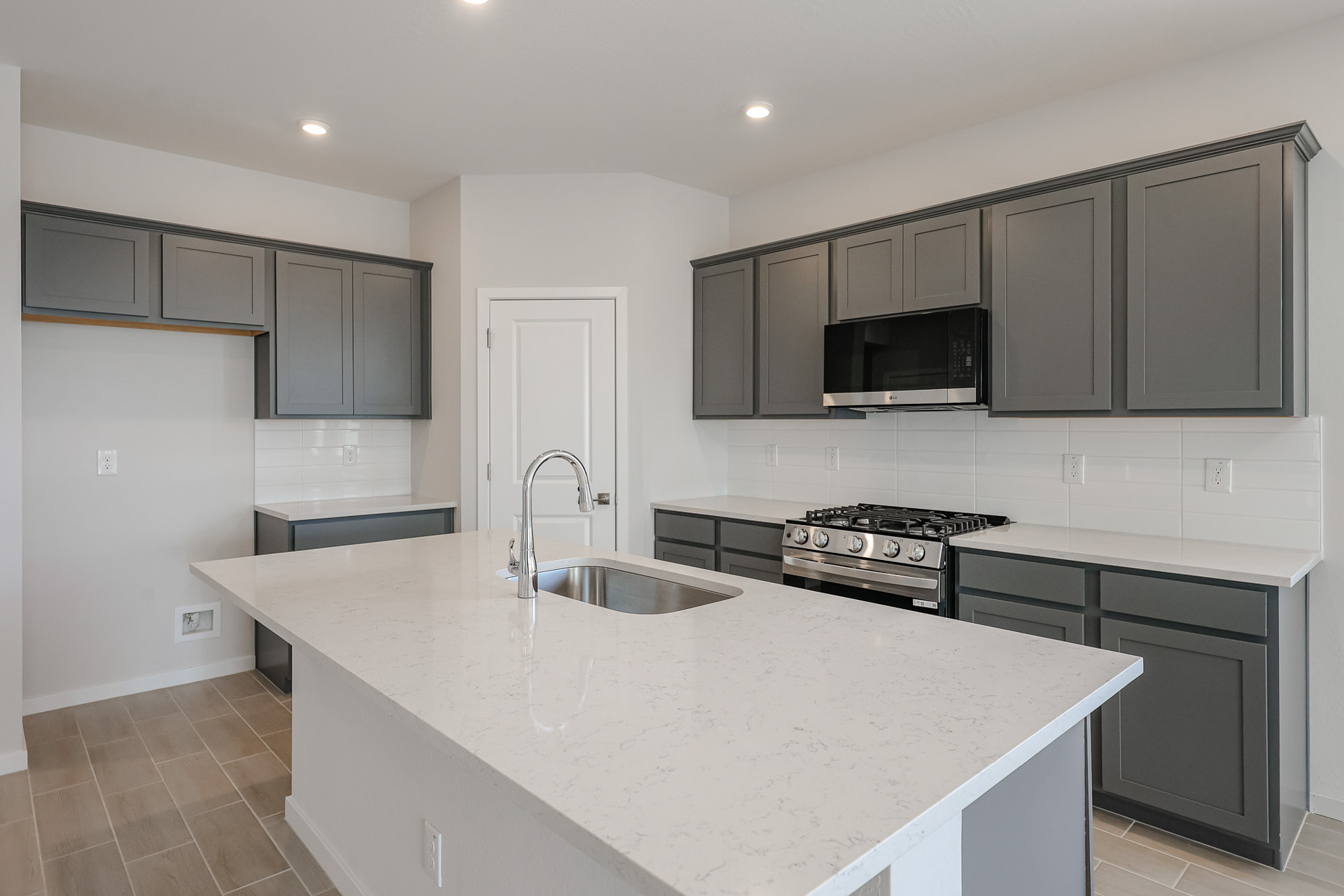Modern kitchen with gray cabinets, white quartz countertops, stainless steel appliances, island sink, gas stove, and white tile backsplash under recessed lighting.