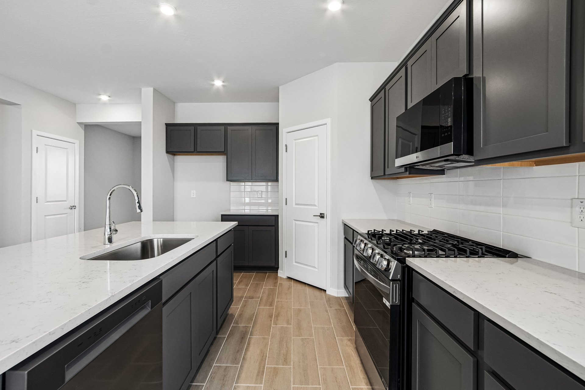 Modern kitchen with black cabinets, stainless steel appliances, white countertops, a sink island, and wood-look tile flooring. The space is brightly lit with recessed ceiling lights.