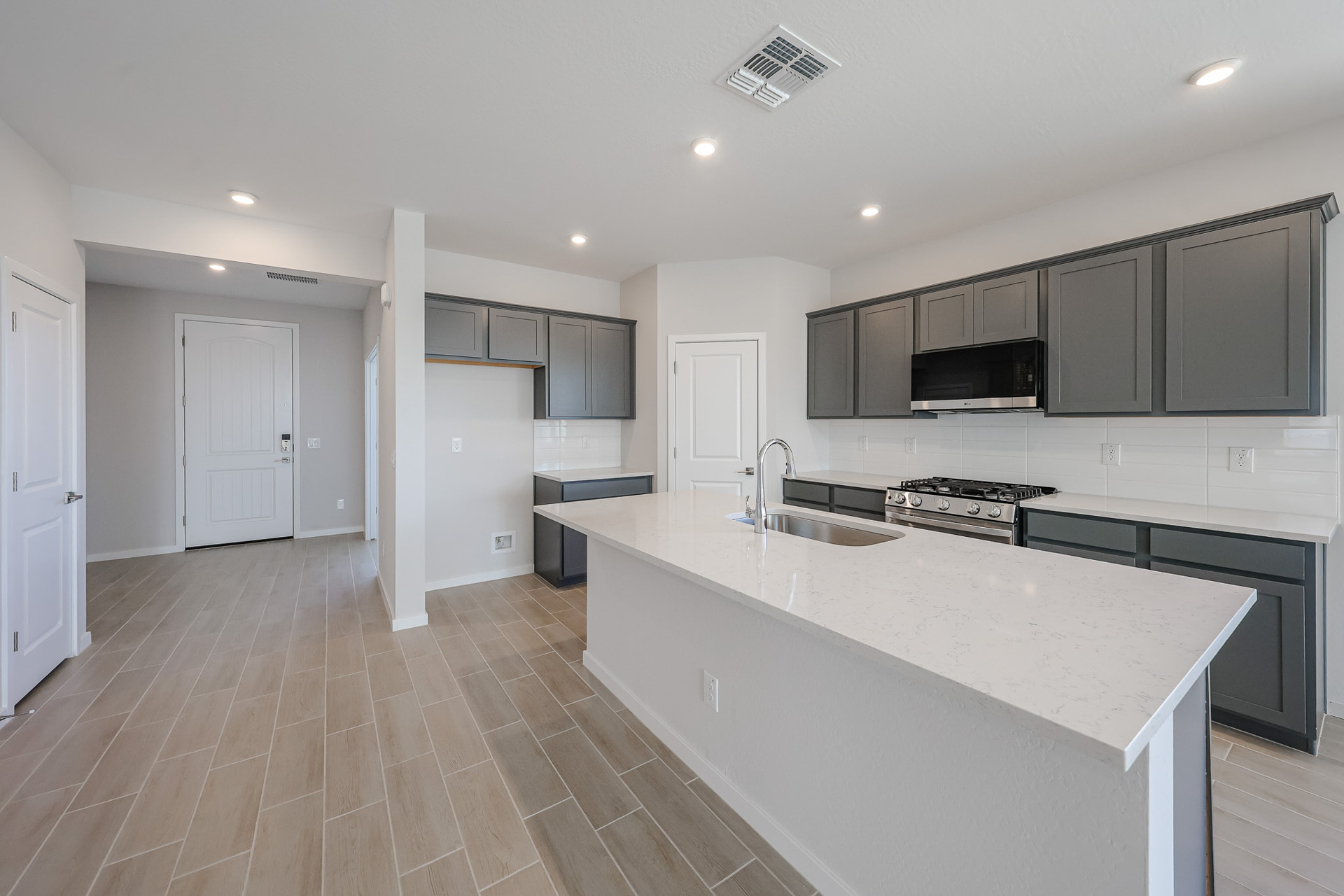 Modern kitchen with gray cabinets, white quartz countertops, stainless steel appliances, tile flooring, and an island with a sink; open layout leading to an entryway and an adjacent room.