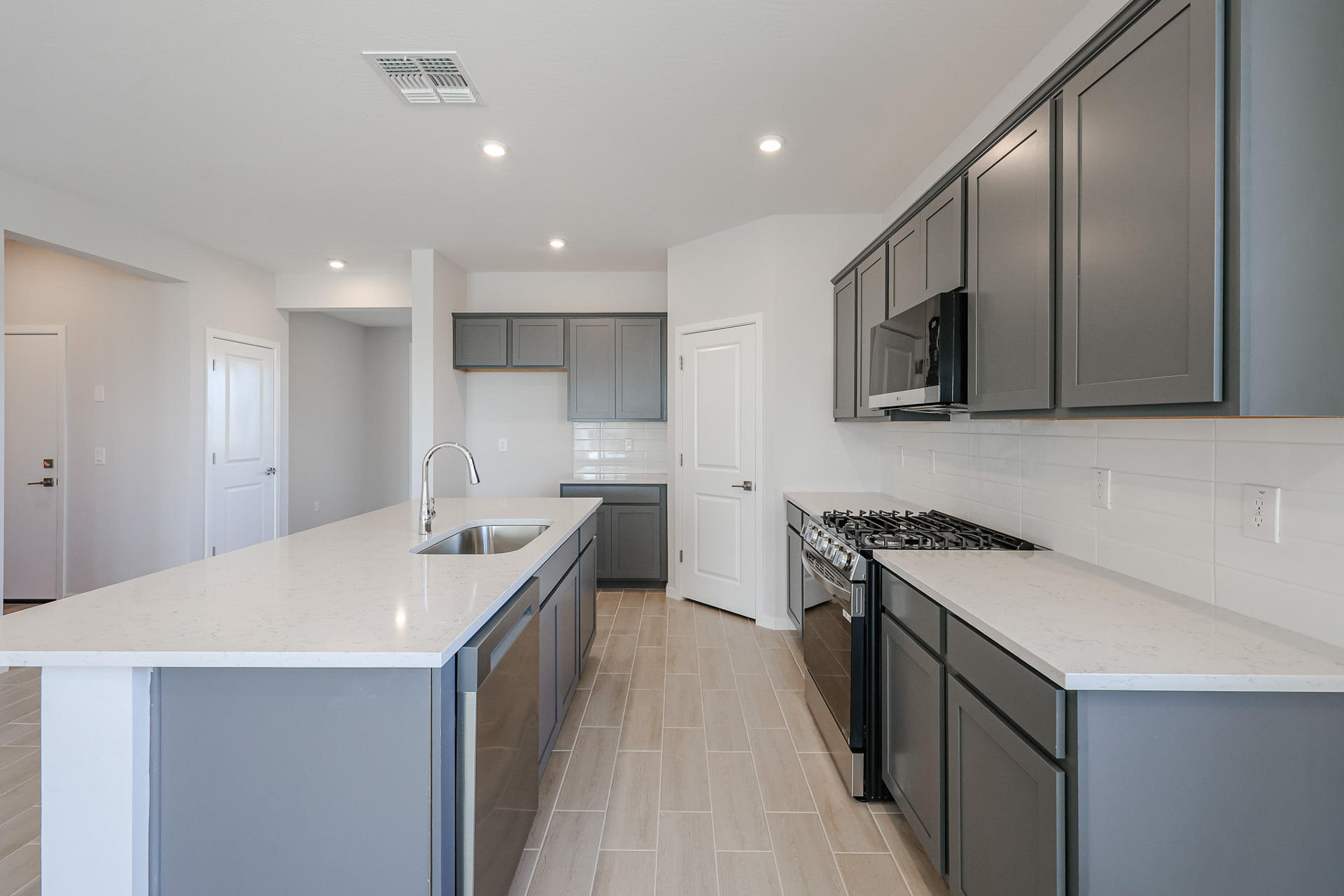 Modern kitchen with gray cabinets, white countertops, a center island with a sink, stainless steel appliances, and light wood flooring. The space is well-lit and minimalistic.