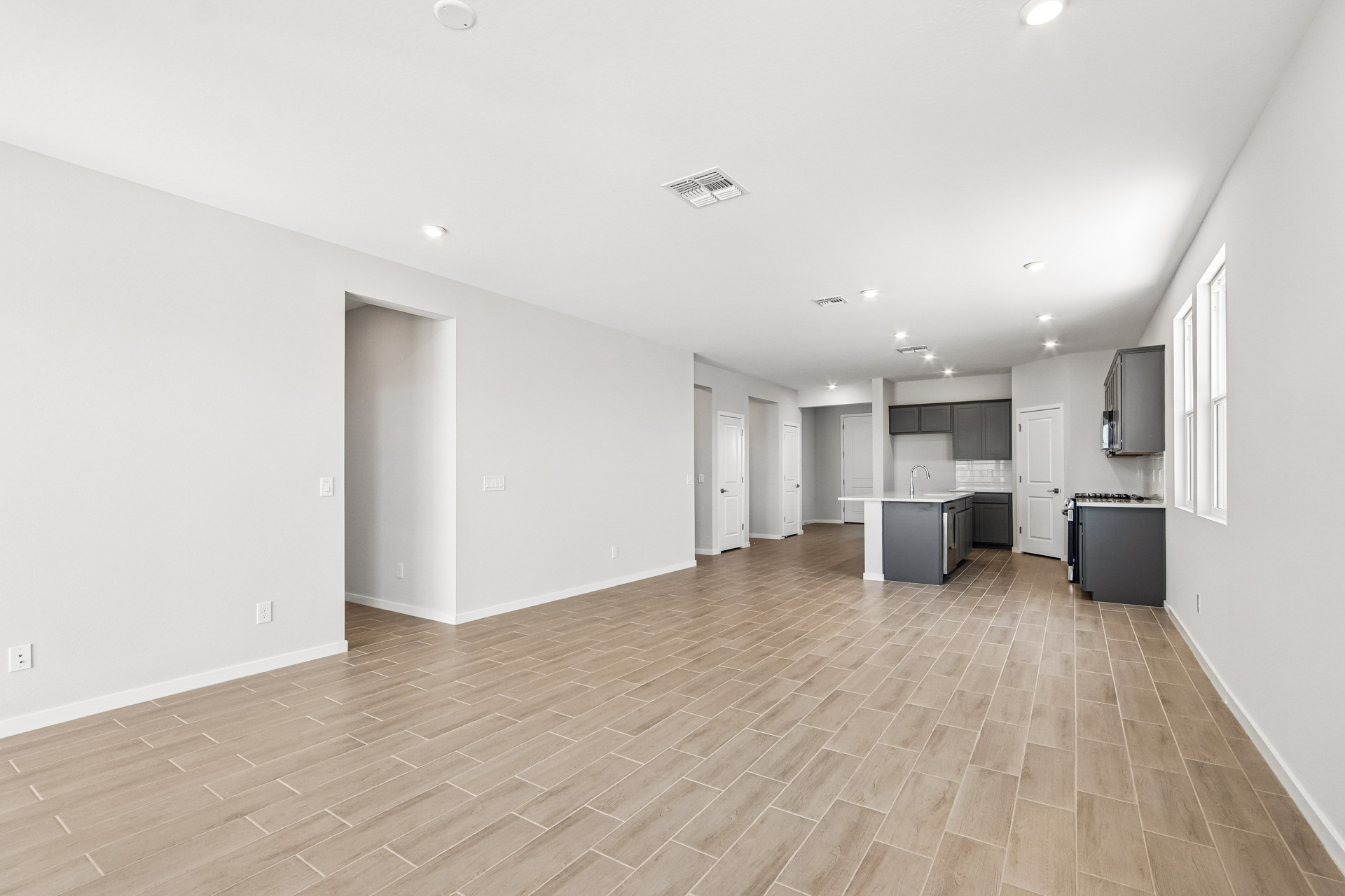 Spacious, empty open-plan room with light brown tile floors, white walls, recessed lighting, and a modern kitchen area with gray cabinets and stainless steel appliances.
