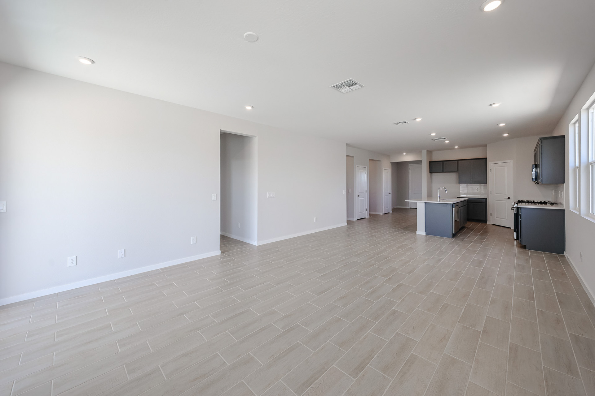 Spacious open-plan living area with light tile flooring, recessed lighting, large windows, and a modern kitchen with gray cabinets and a center island in the background.
