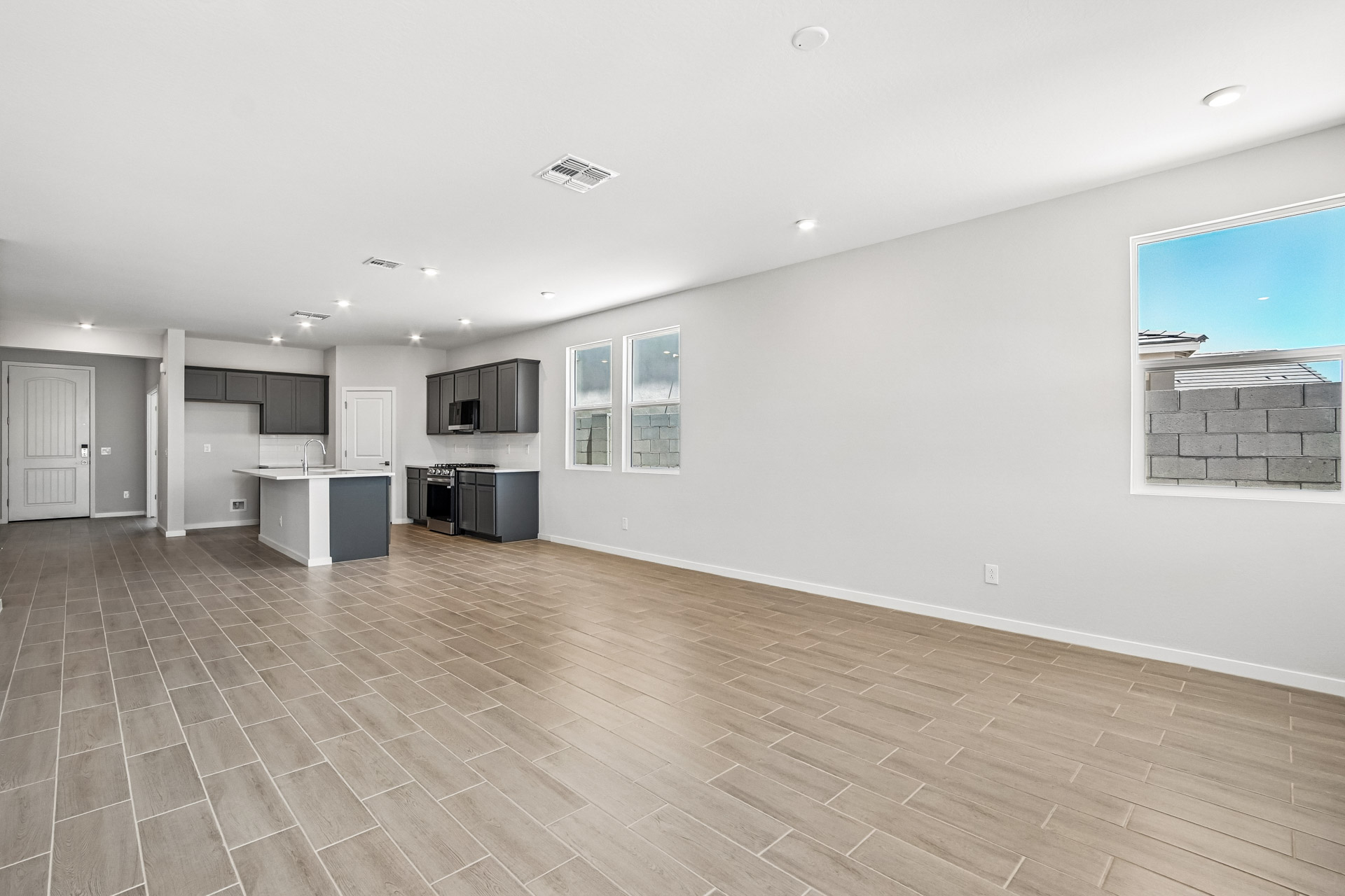 Spacious, unfurnished open-plan room with tile flooring, white walls, large windows, and a modern kitchen area with grey cabinets in the background.