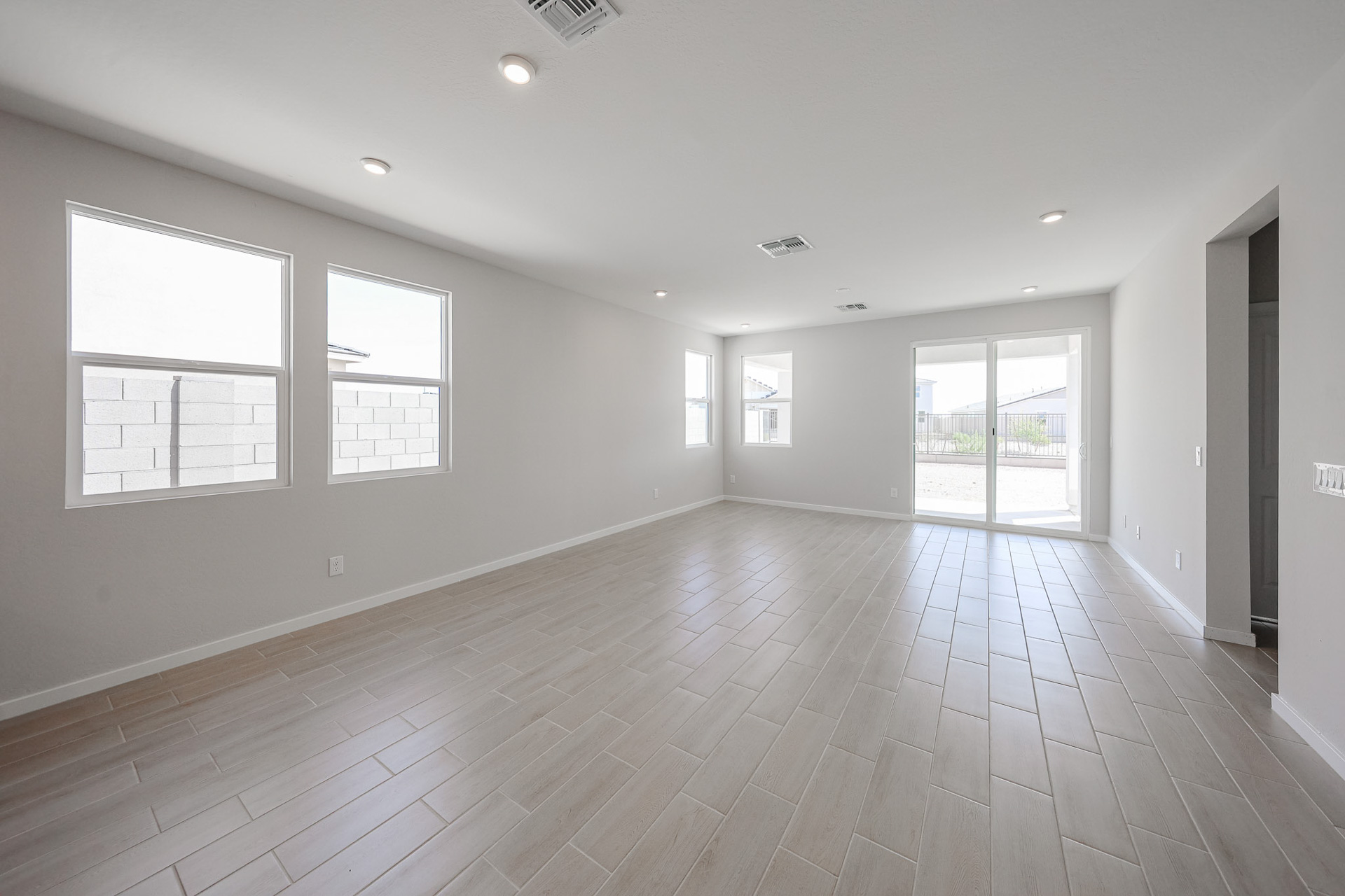Spacious empty room with large tile floor, white walls, recessed lighting, two windows, and sliding glass doors leading outside.