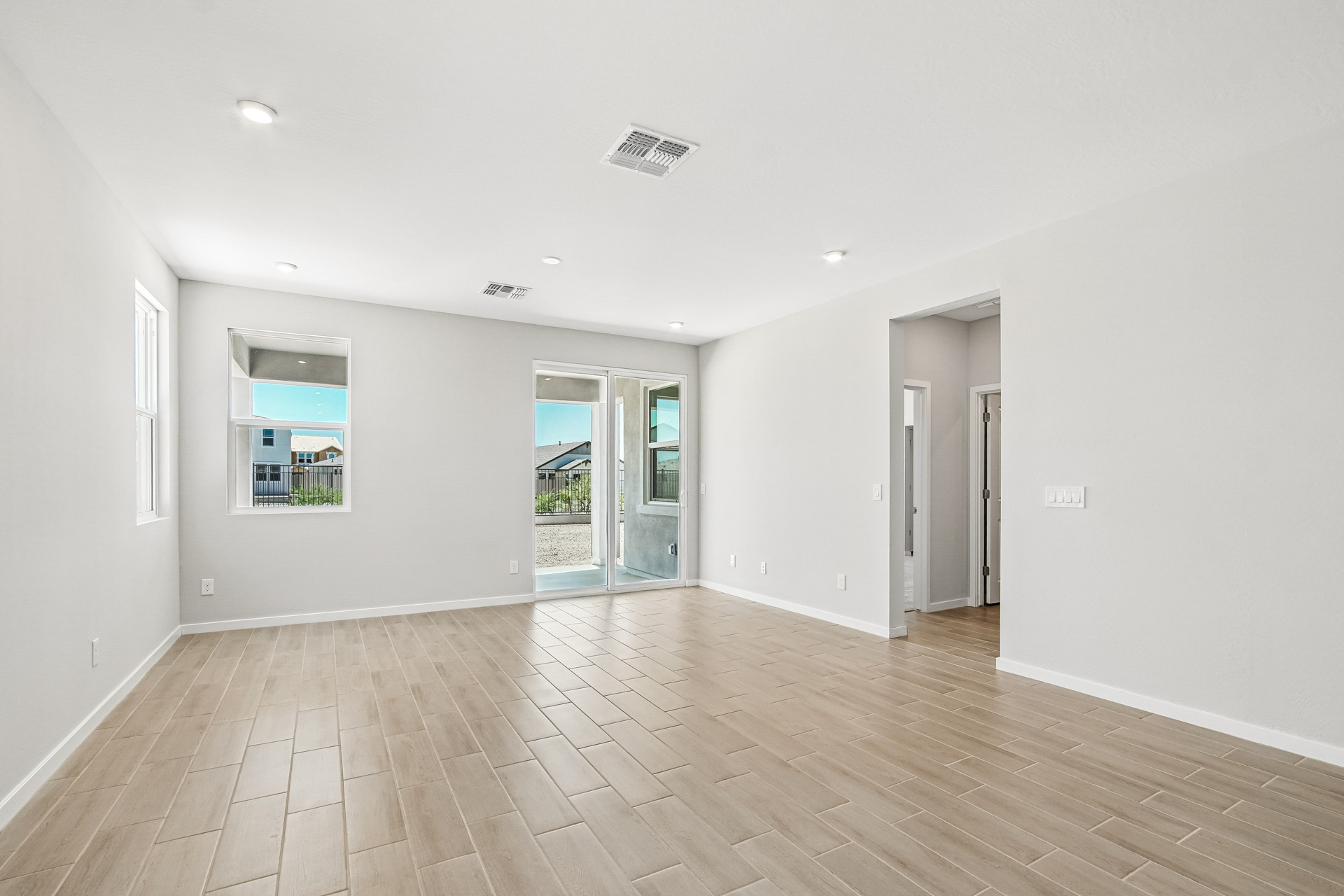 Empty living room with light wood-look tile flooring, white walls, recessed lighting, and large windows with a glass door leading to a covered patio.