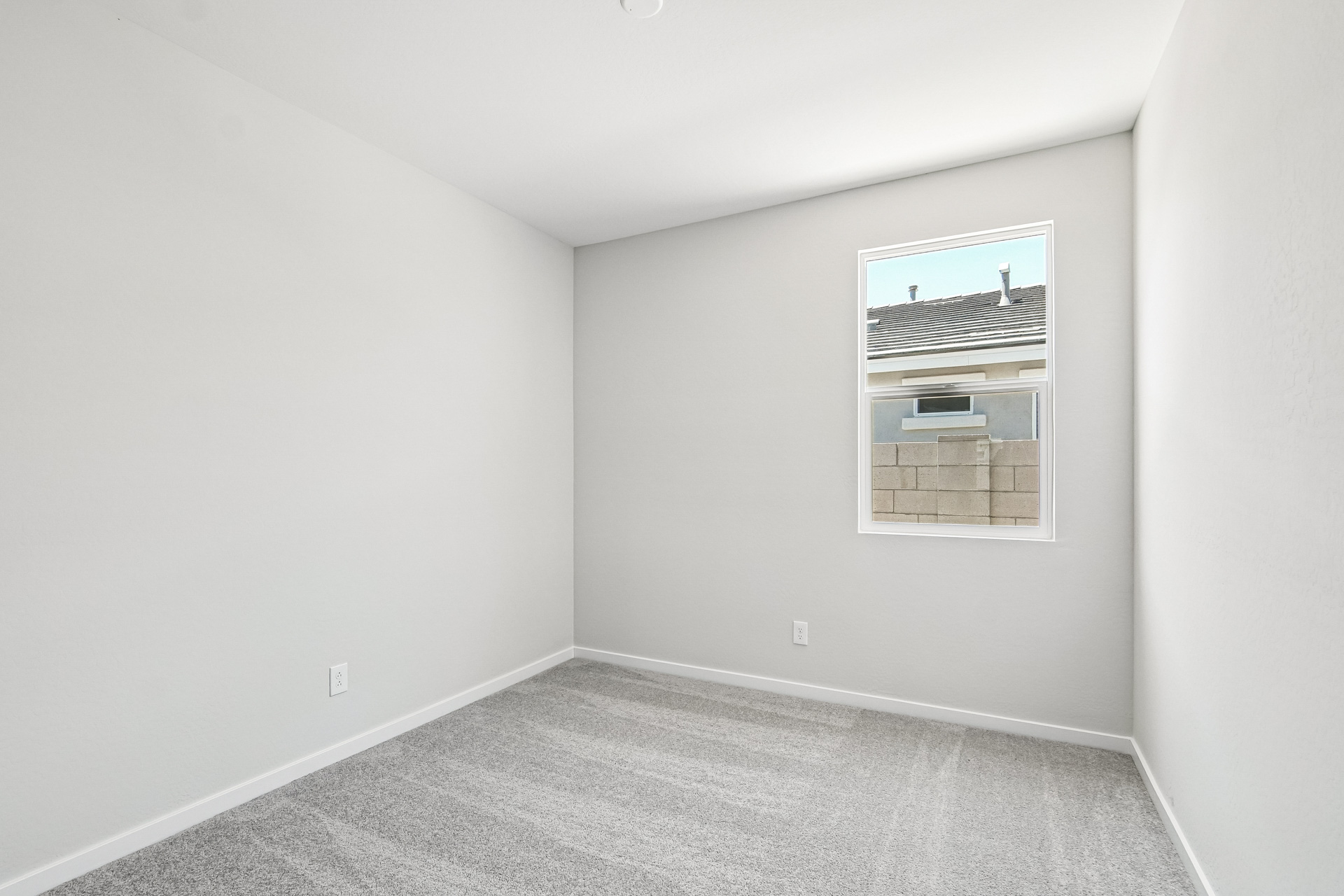 Empty room with light gray carpet, white walls, and a single window showing a partial view of a roof and fence outside.