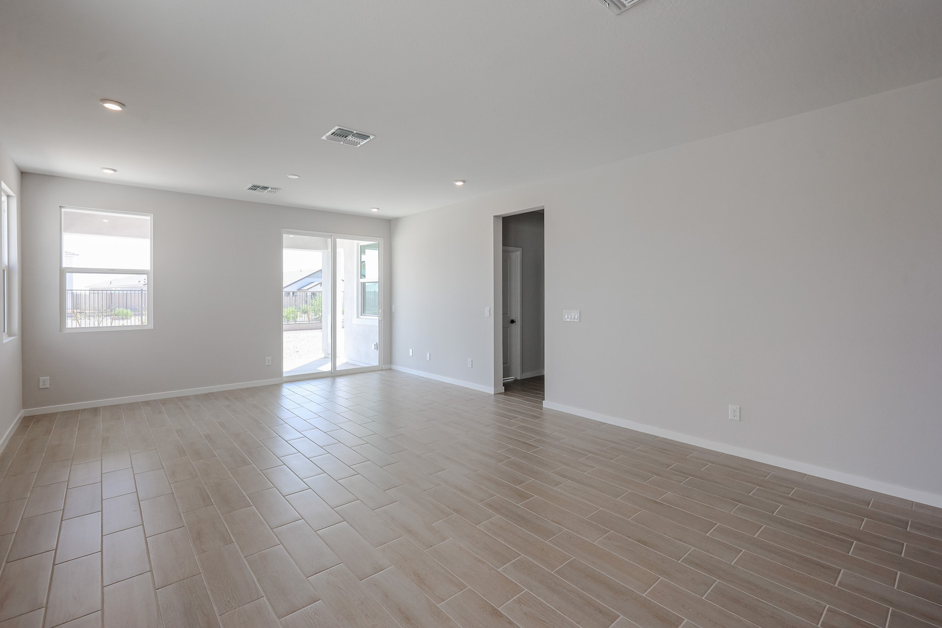 Empty, unfurnished room with light-gray walls, tan wood-look tile flooring, recessed ceiling lights, and glass doors leading outside. Two windows provide additional natural light.